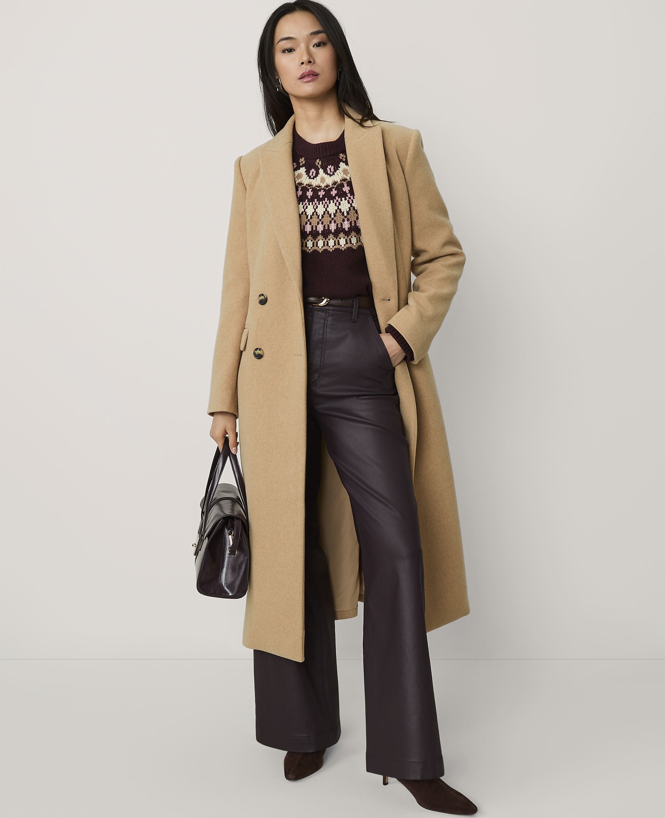 A woman wearing a tan coat, patterned sweater, and dark leather trousers stands against a plain white background.