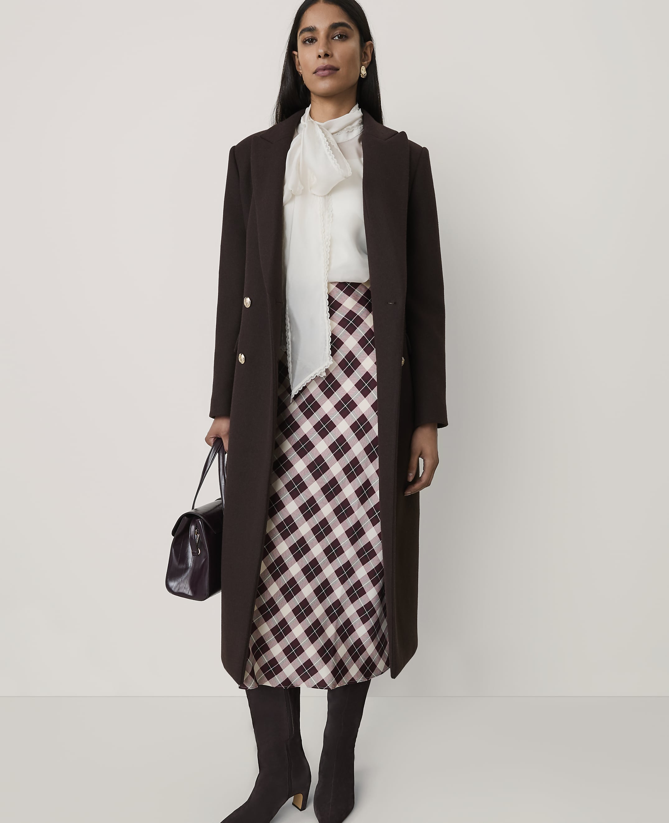 A woman wearing a long brown coat, a white blouse, and a plaid skirt stands against a plain, light-colored background.