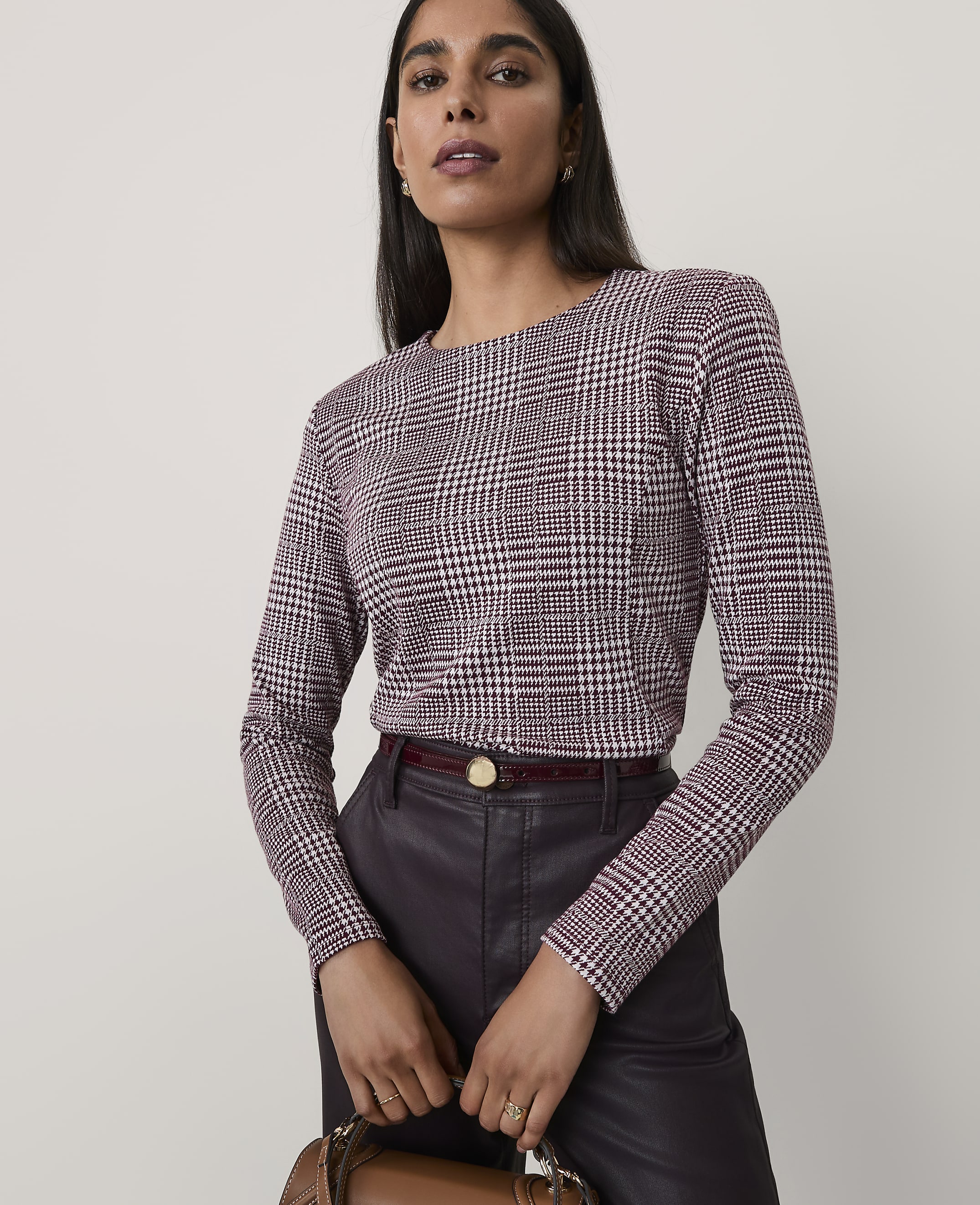 A woman wearing a maroon and white houndstooth top and dark leather trousers stands against a plain, light-colored background.