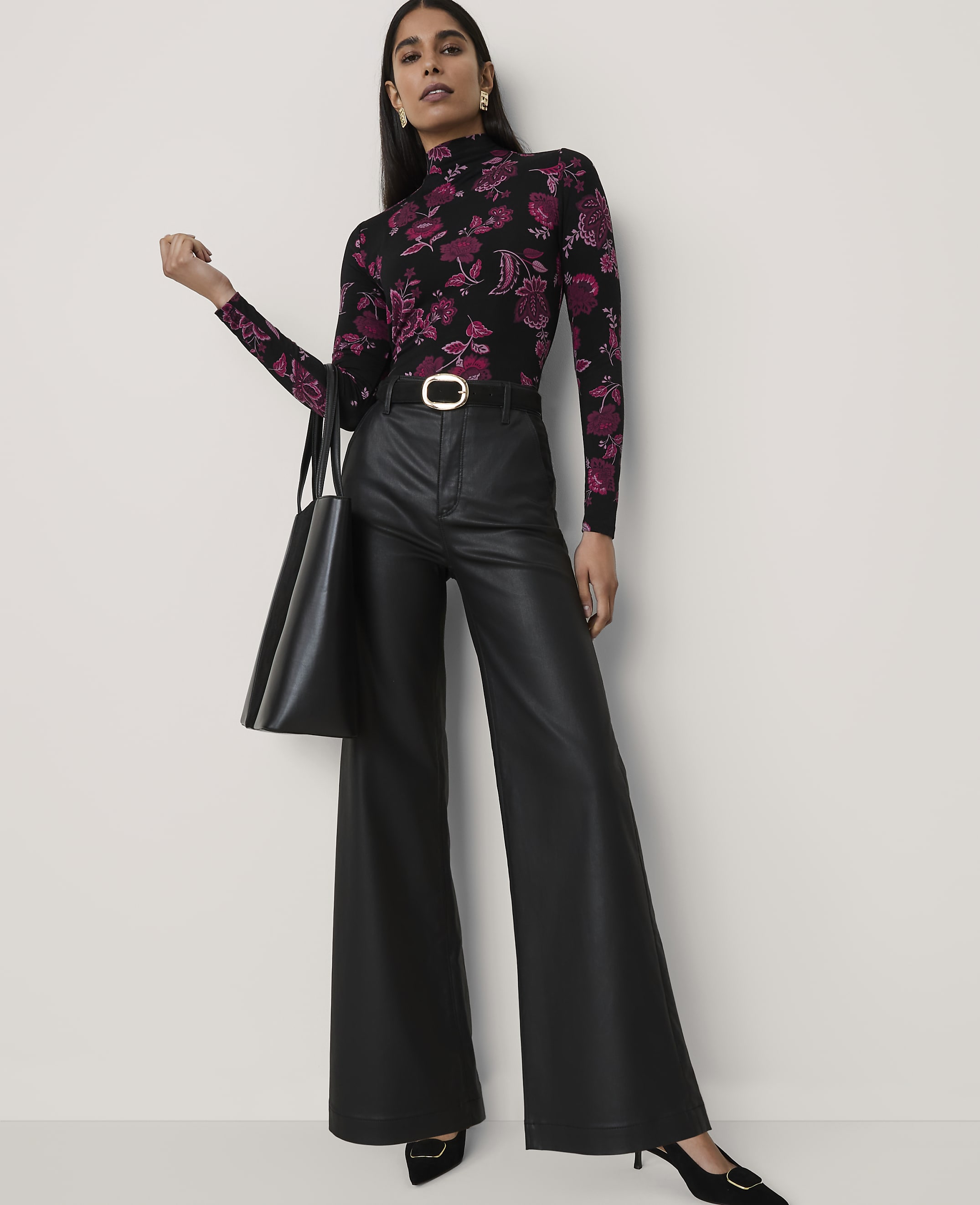 A woman wearing a floral black and purple turtleneck and wide-leg black leather trousers stands against a plain light gray background.
