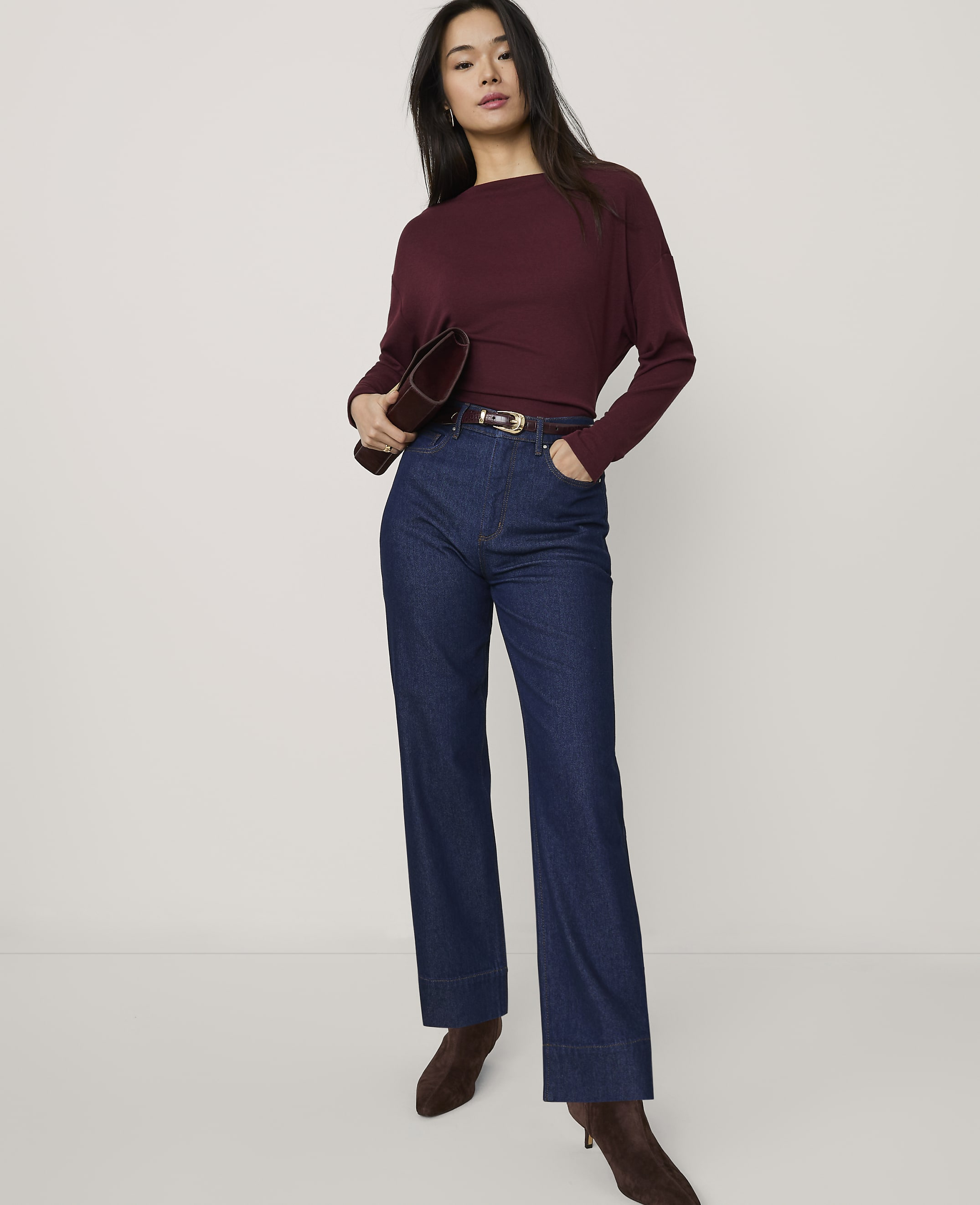 A woman wearing a burgundy top and wide-leg blue jeans stands against a plain white background.