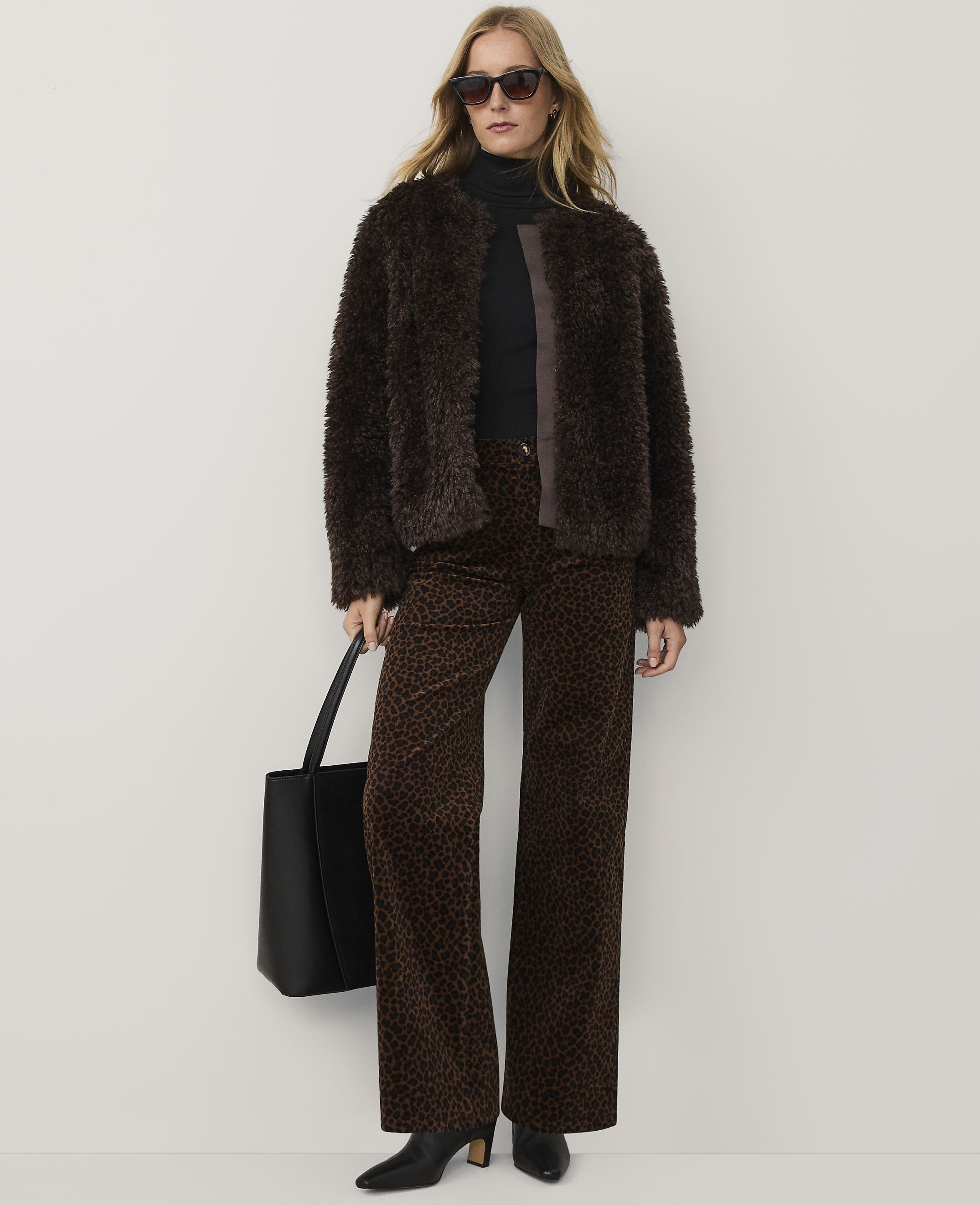 A woman wearing a brown faux-fur jacket, leopard print trousers, and sunglasses stands against a plain white background.