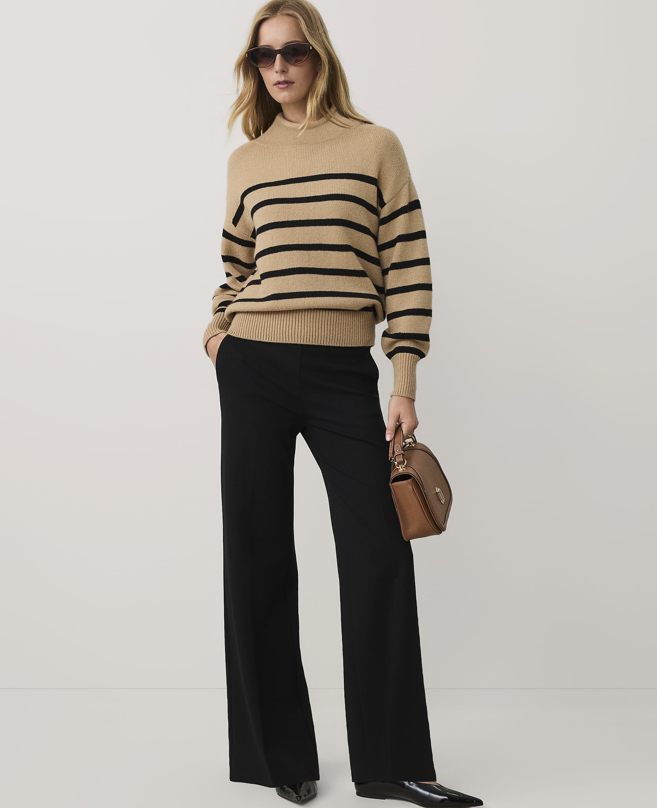 A woman wearing a tan striped sweater, black wide-leg trousers, and sunglasses stands against a plain white background.