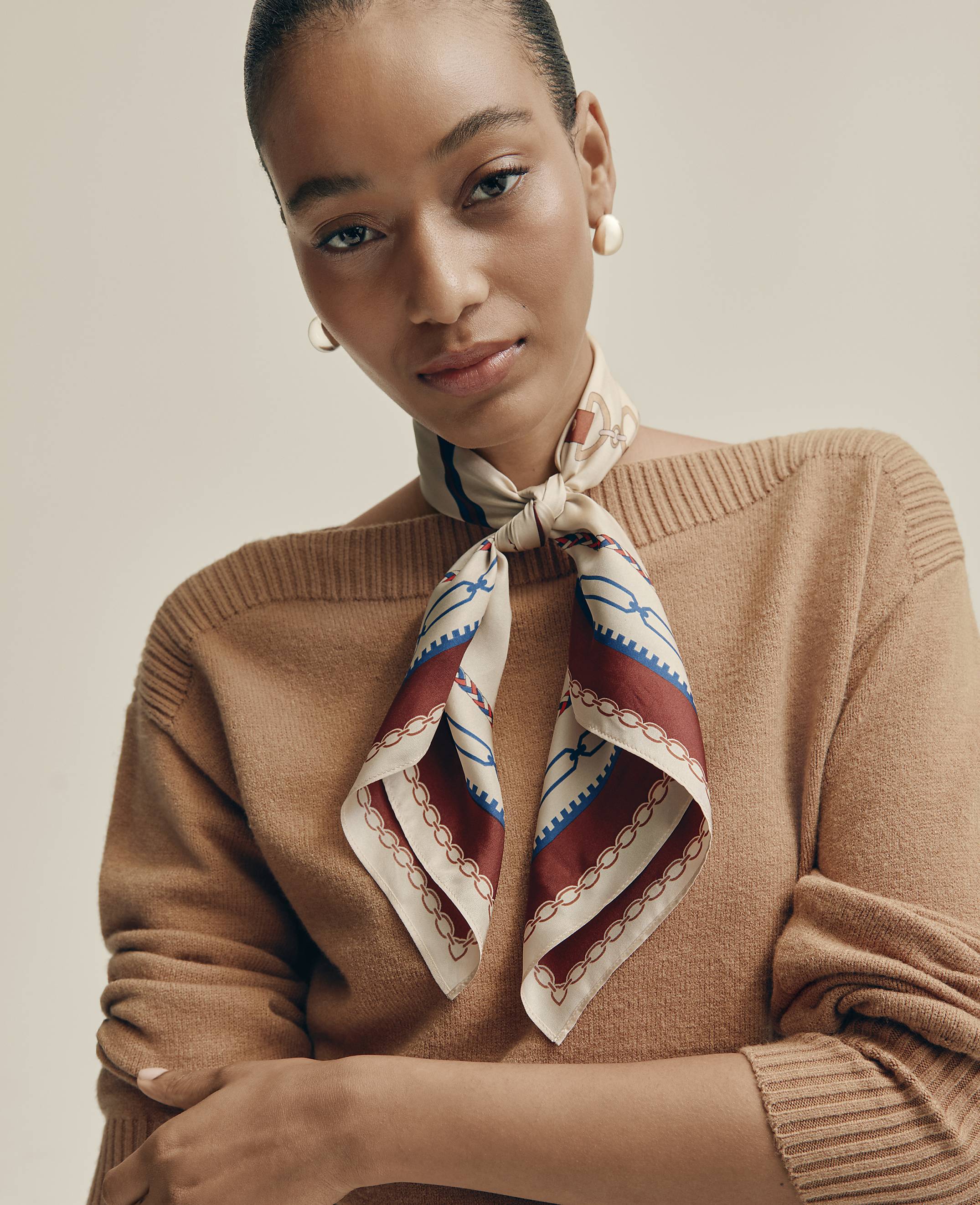 A woman wearing a brown sweater and a patterned silk scarf poses against a plain, neutral-toned background.