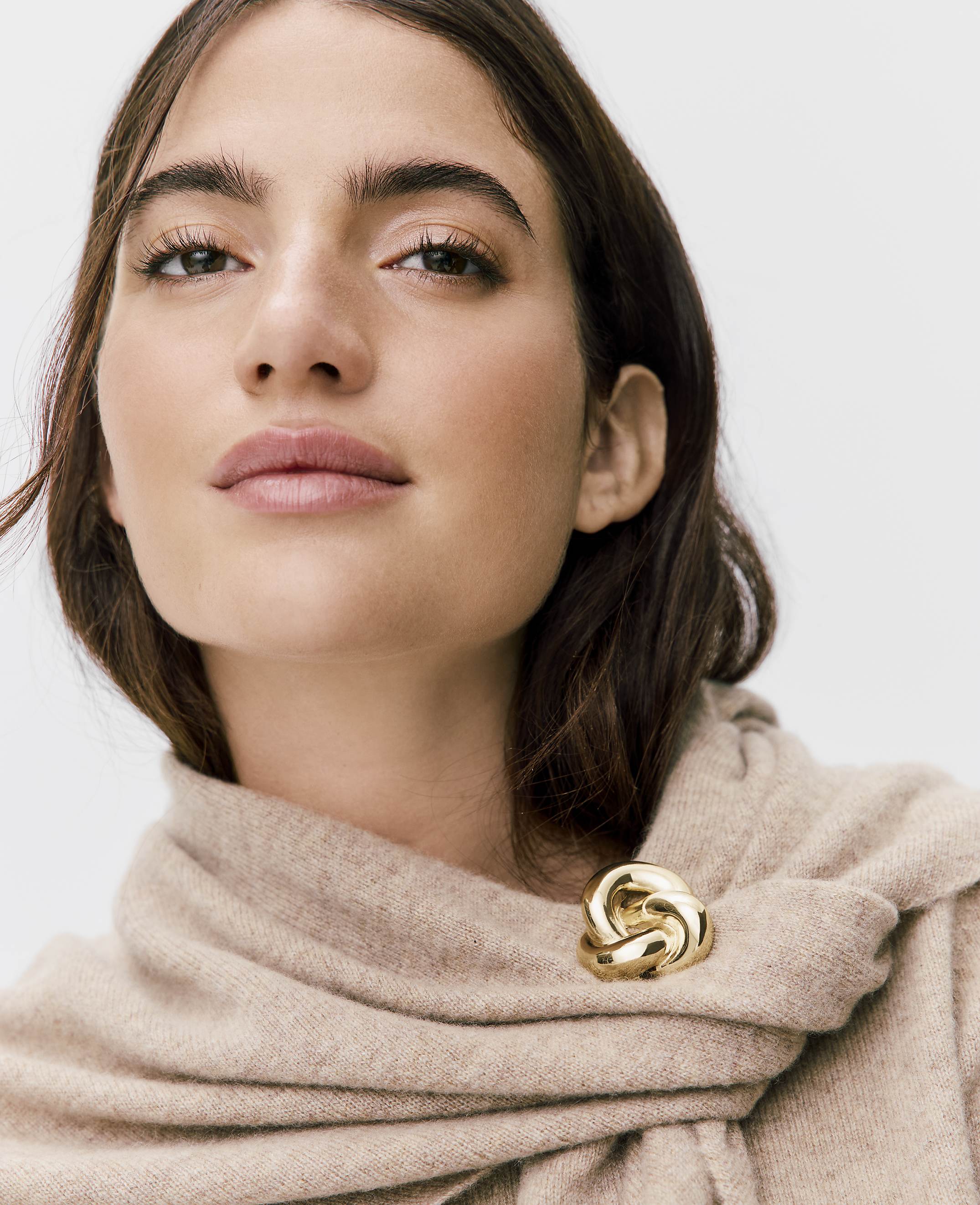 A woman with brown hair and a beige scarf fastened with a gold brooch poses against a plain white background.