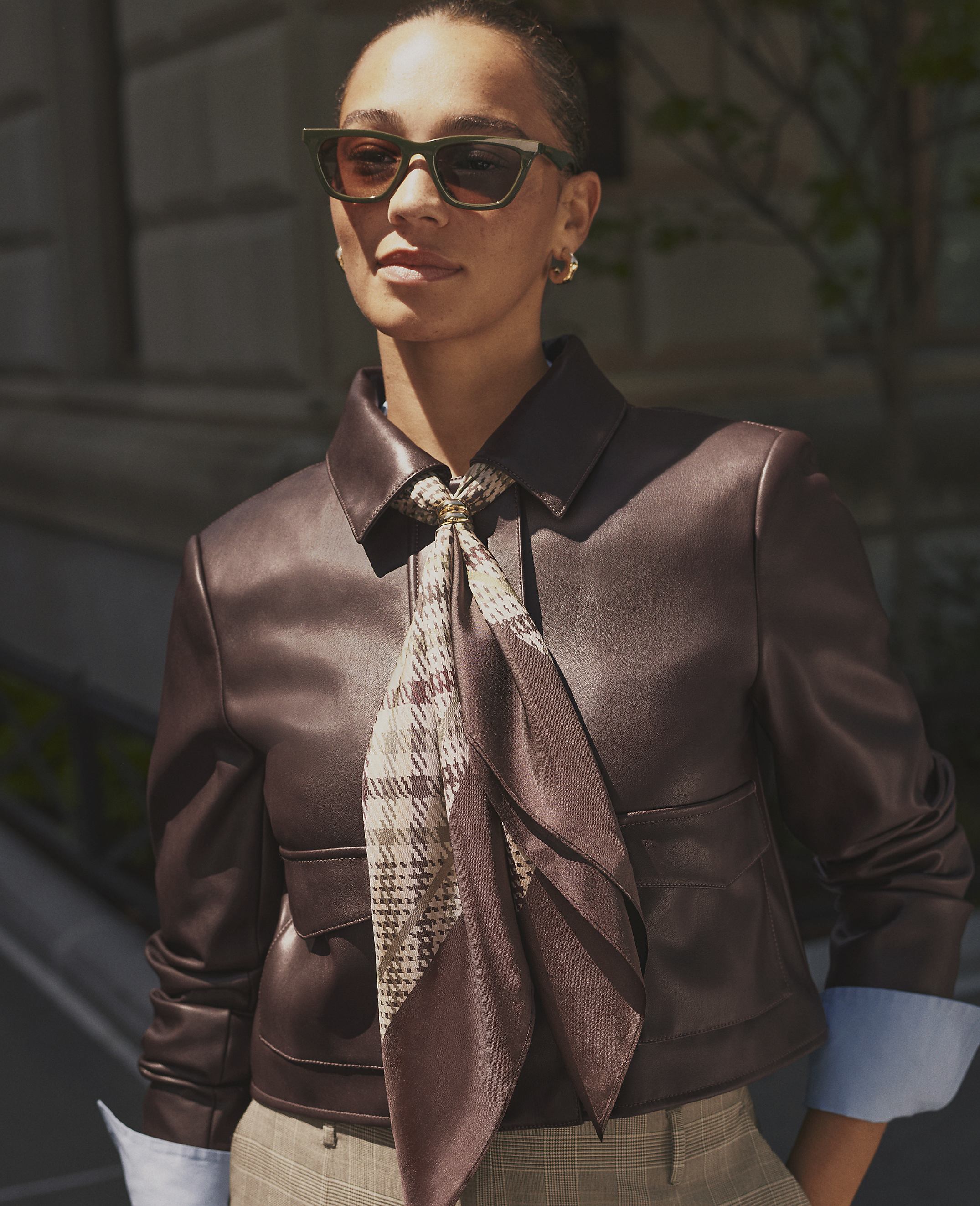 A woman wearing sunglasses, a brown leather jacket, and a patterned scarf stands before a blurred urban building background.