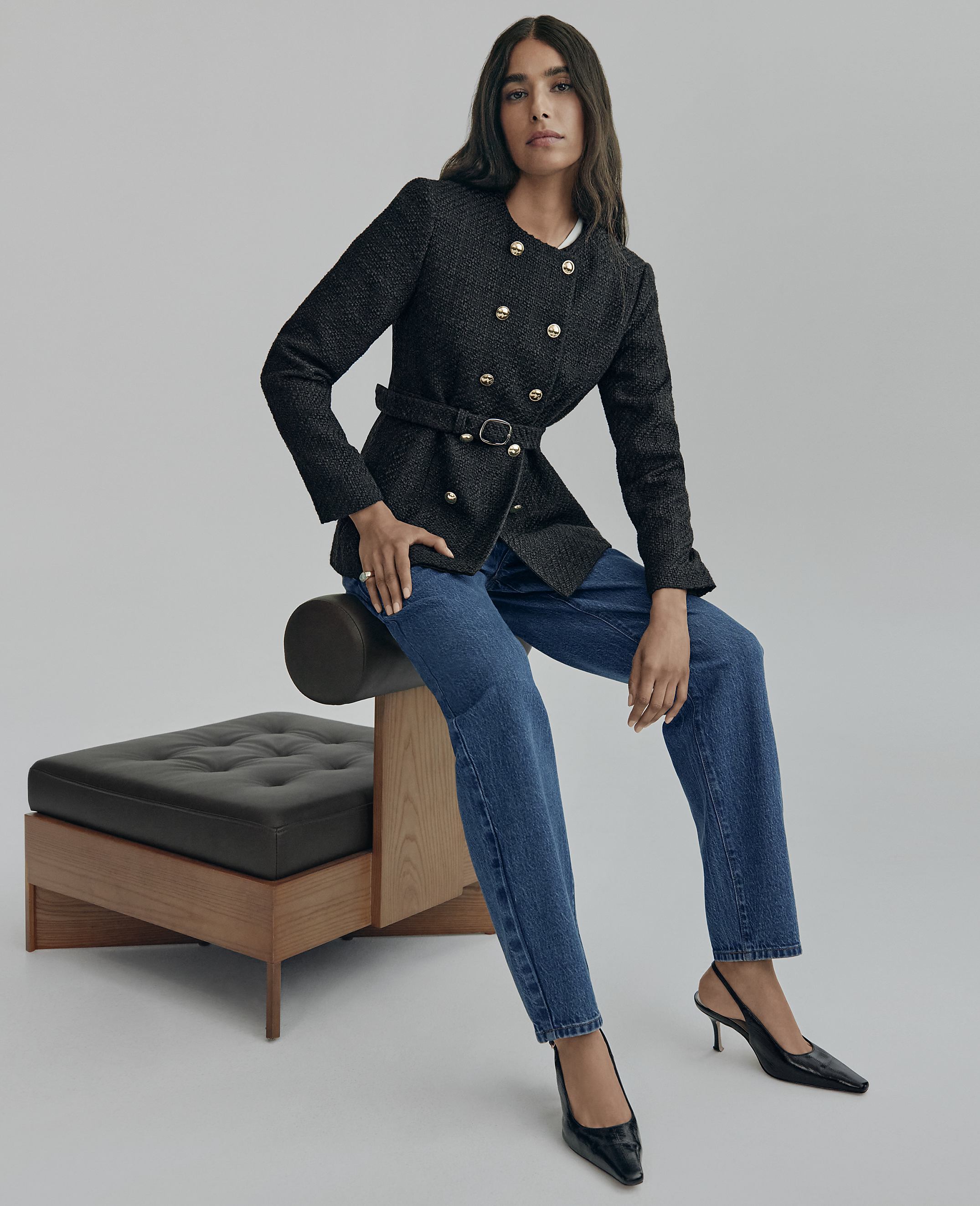 A woman wearing a black double-breasted blazer and blue jeans sits on a modern black leather and wood bench against a plain grey background.