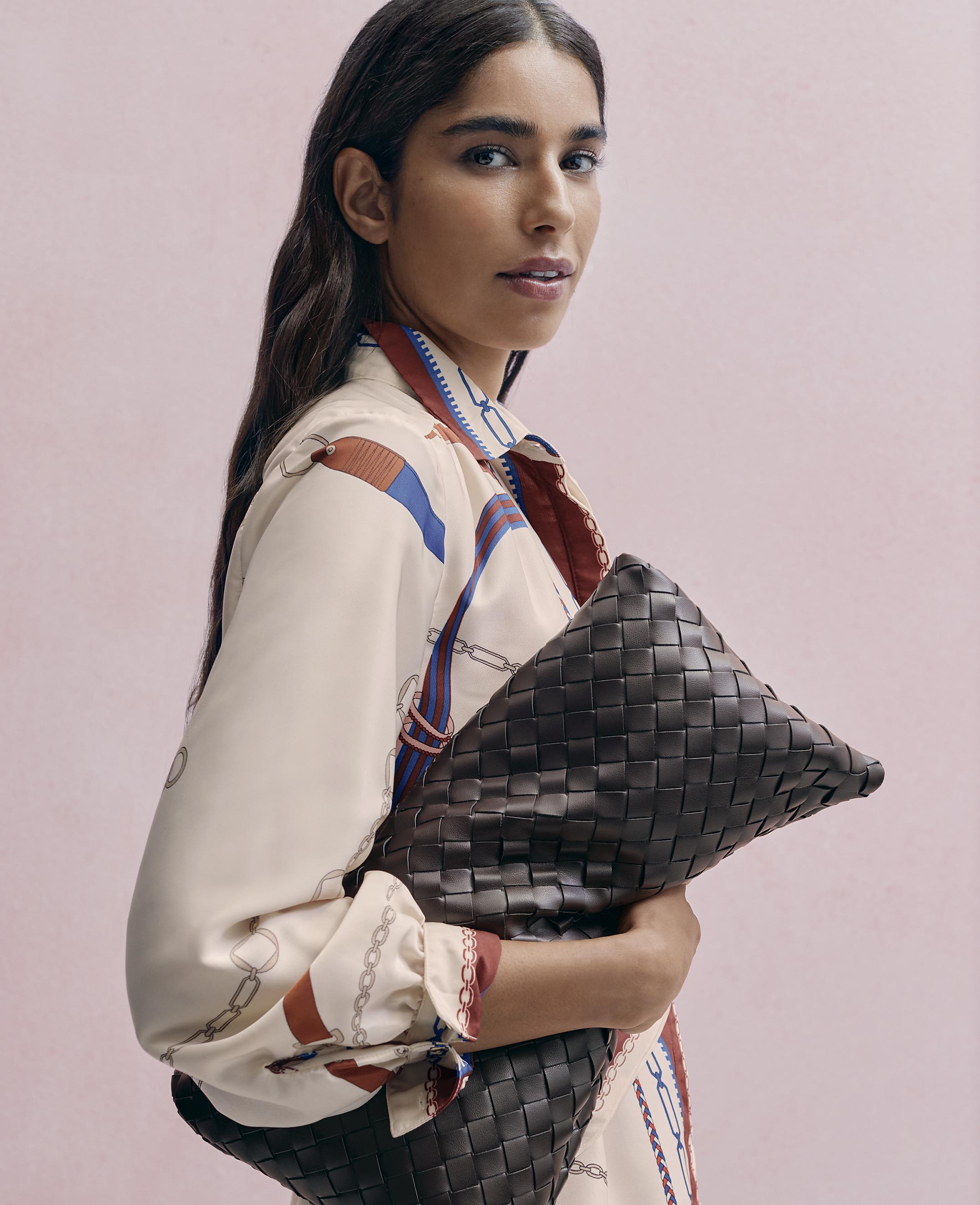 A woman in a patterned cream blouse holds a large woven brown leather clutch against a solid pink background.