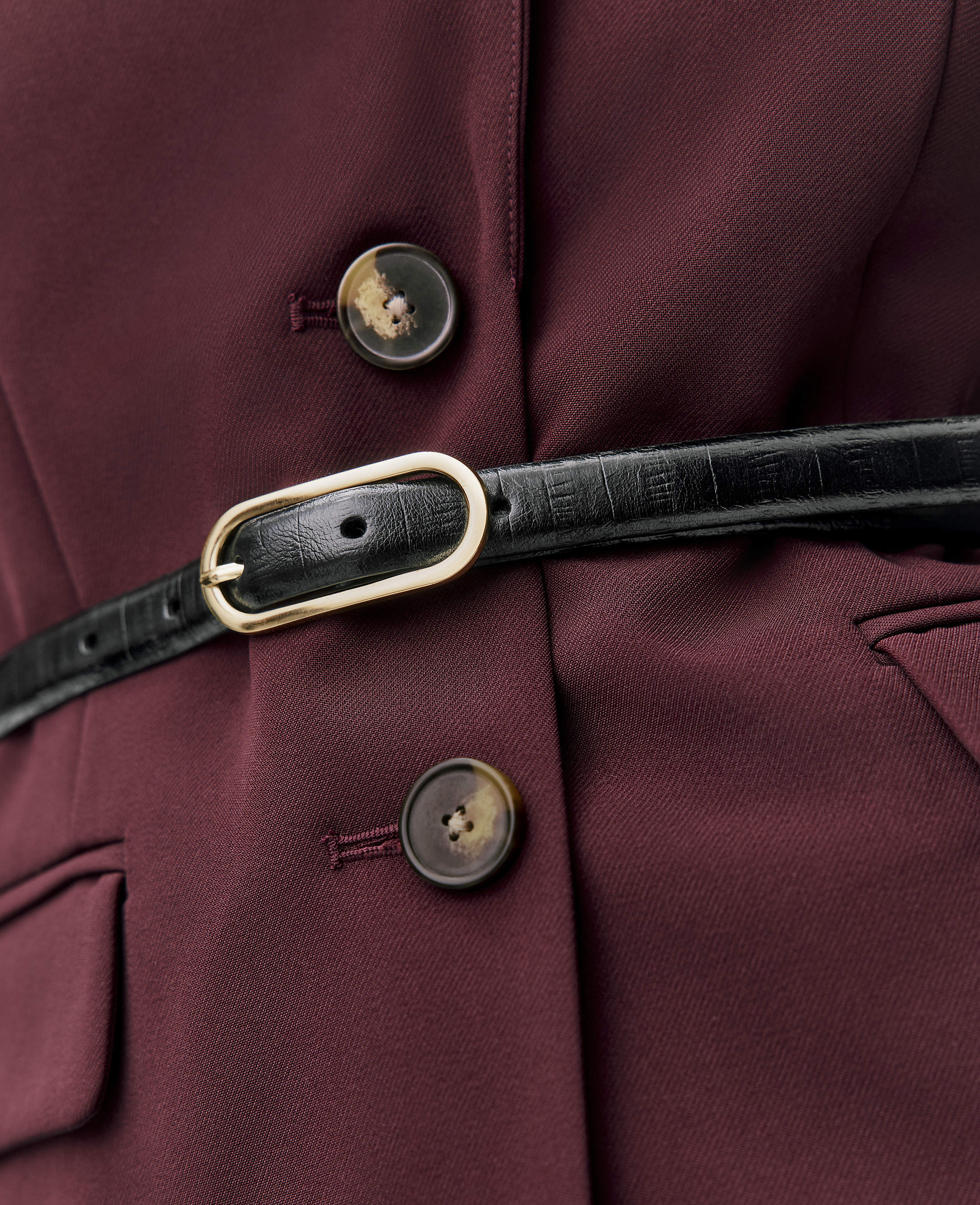 A black leather belt with a gold buckle is fastened across a maroon blazer featuring two brown buttons.