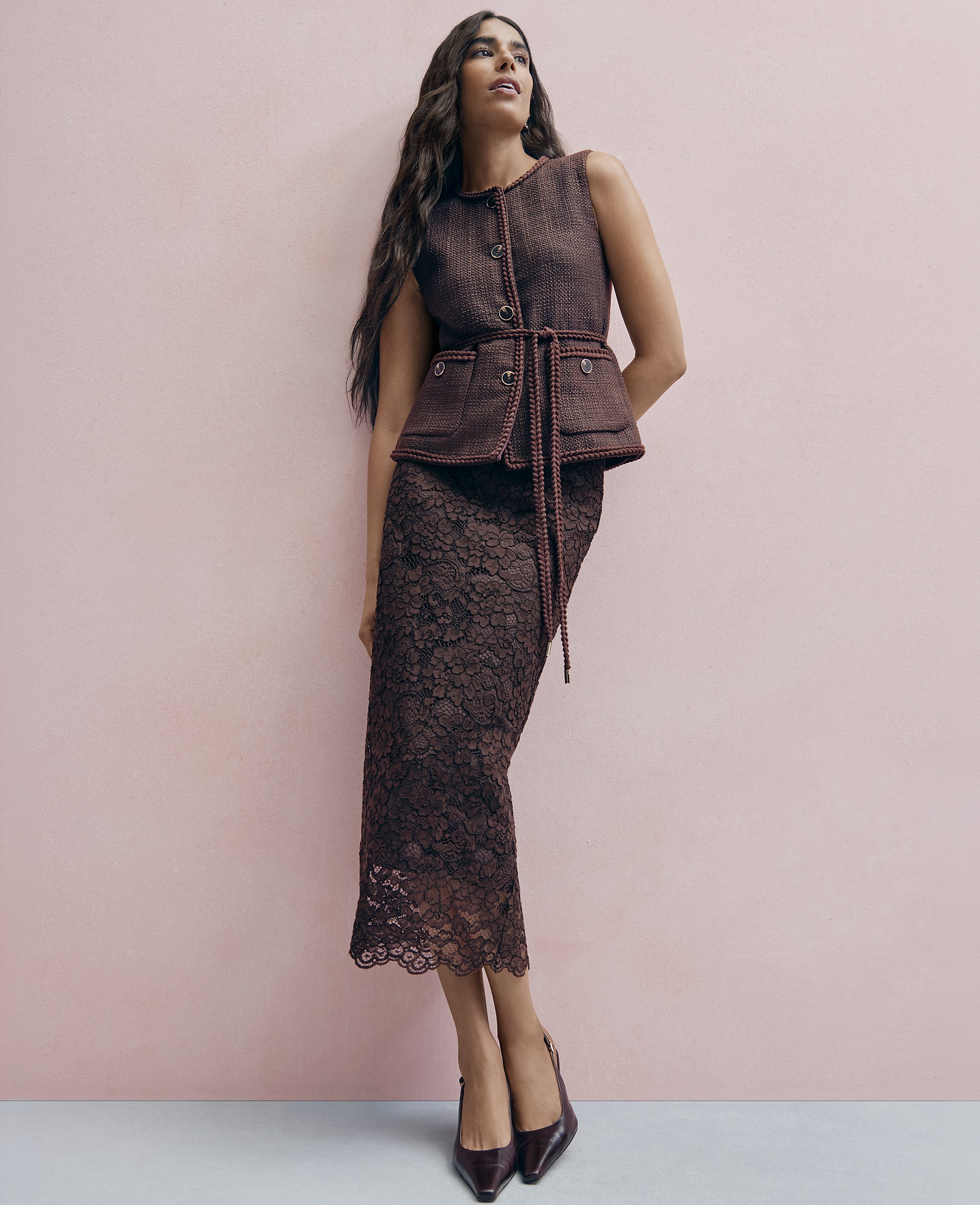 A woman wearing a brown tweed vest and lace skirt stands against a solid pink wall.