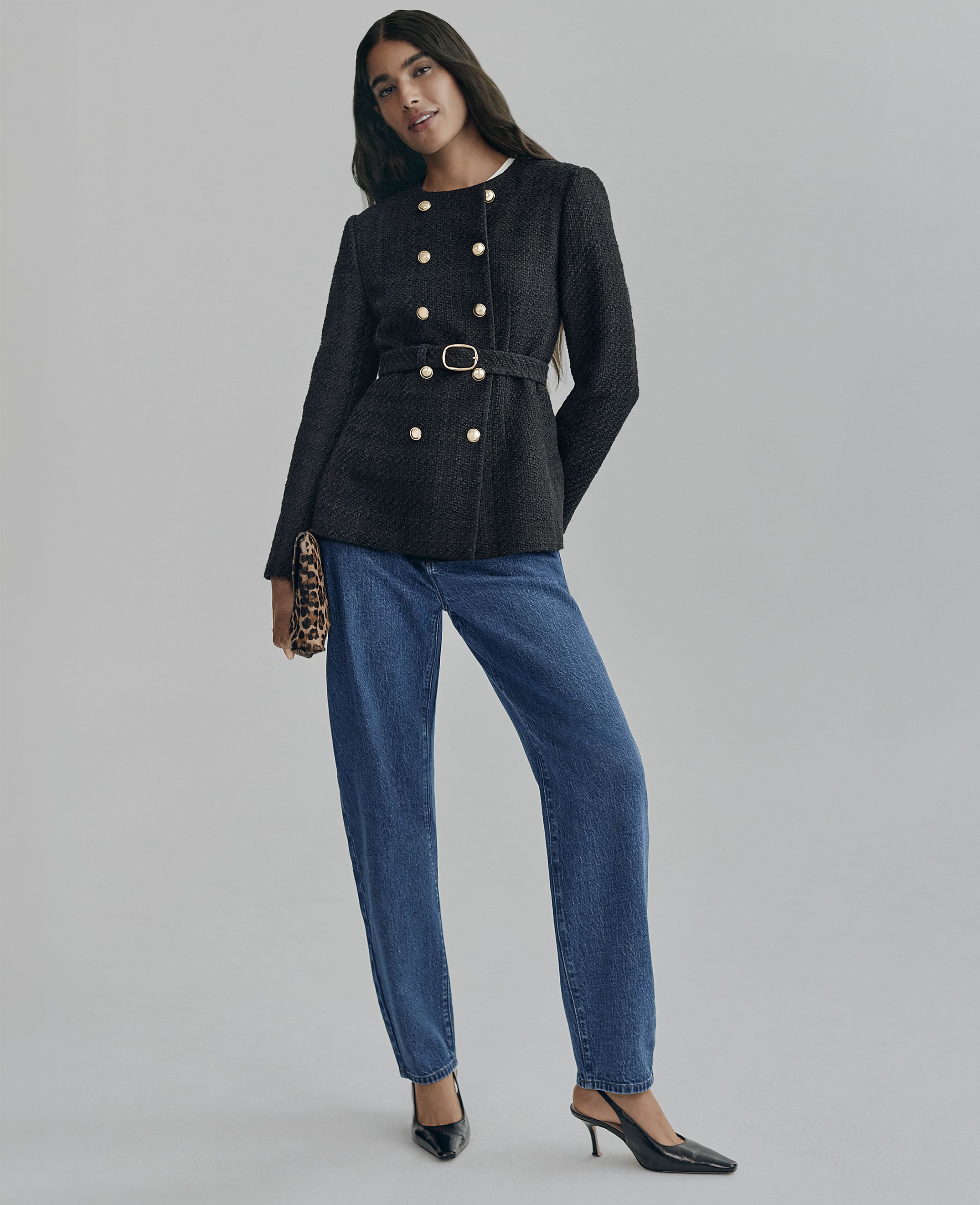 A woman wearing a belted black double-breasted jacket and blue jeans stands against a plain grey background.