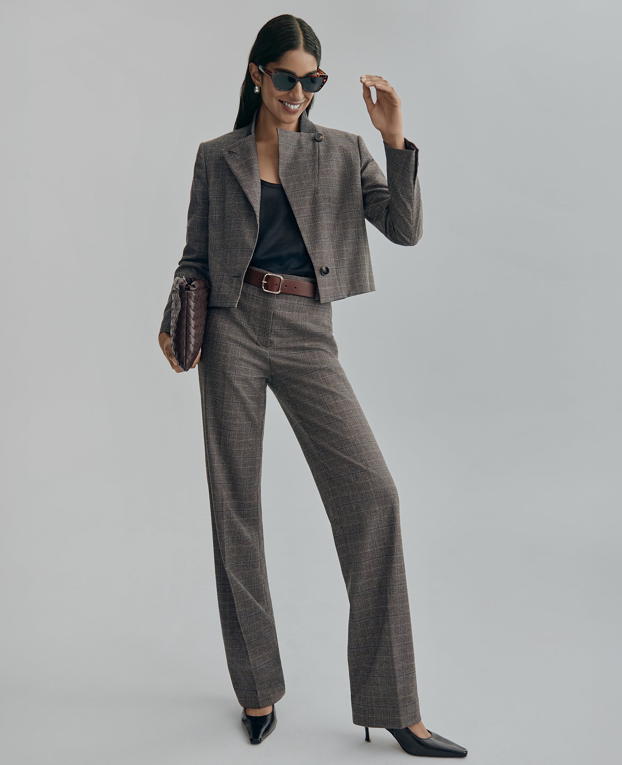 A woman wearing a grey plaid cropped blazer and matching trousers stands against a plain light grey background.