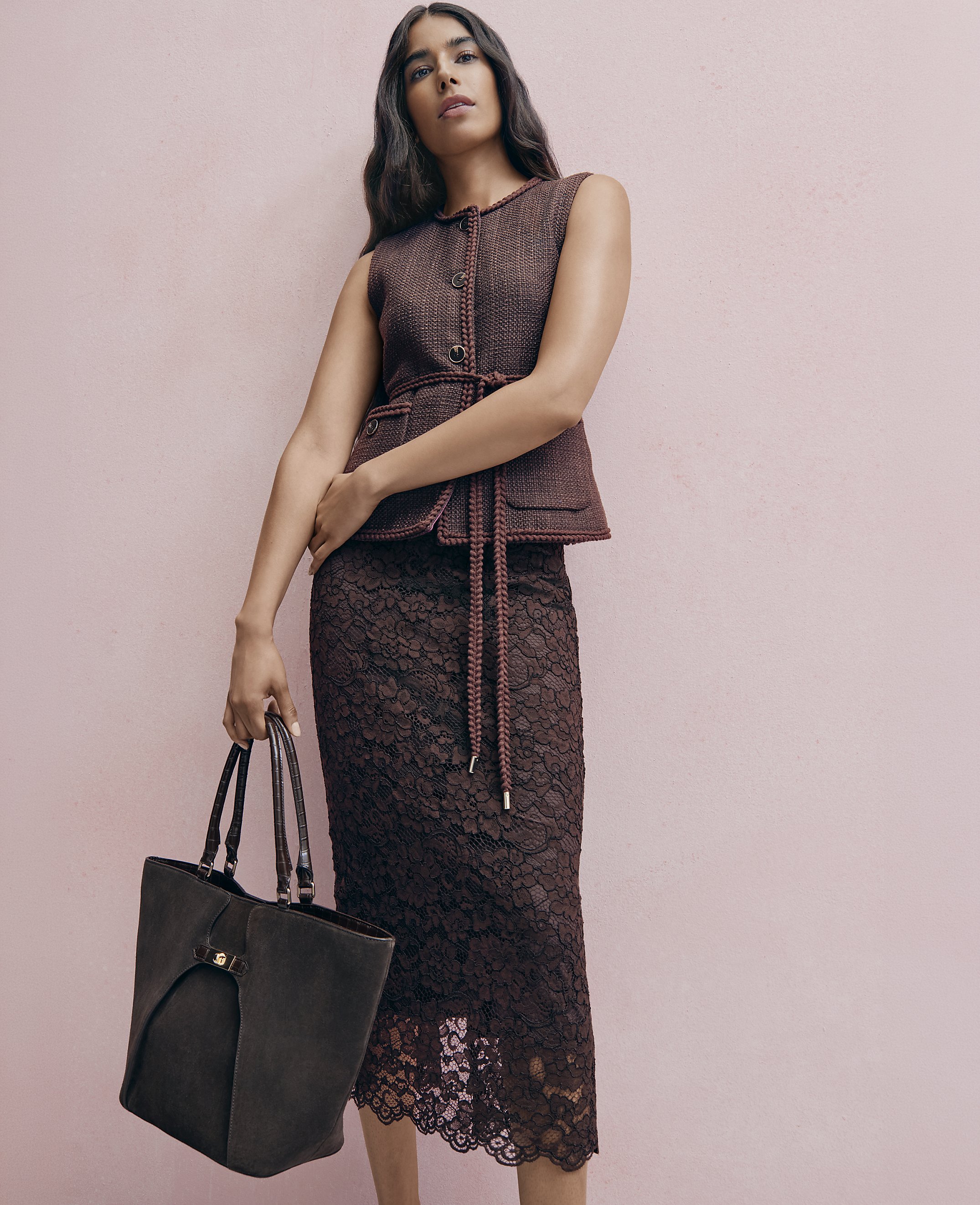 A woman wearing a maroon textured vest and lace skirt holds a large dark tote bag against a plain pink background.