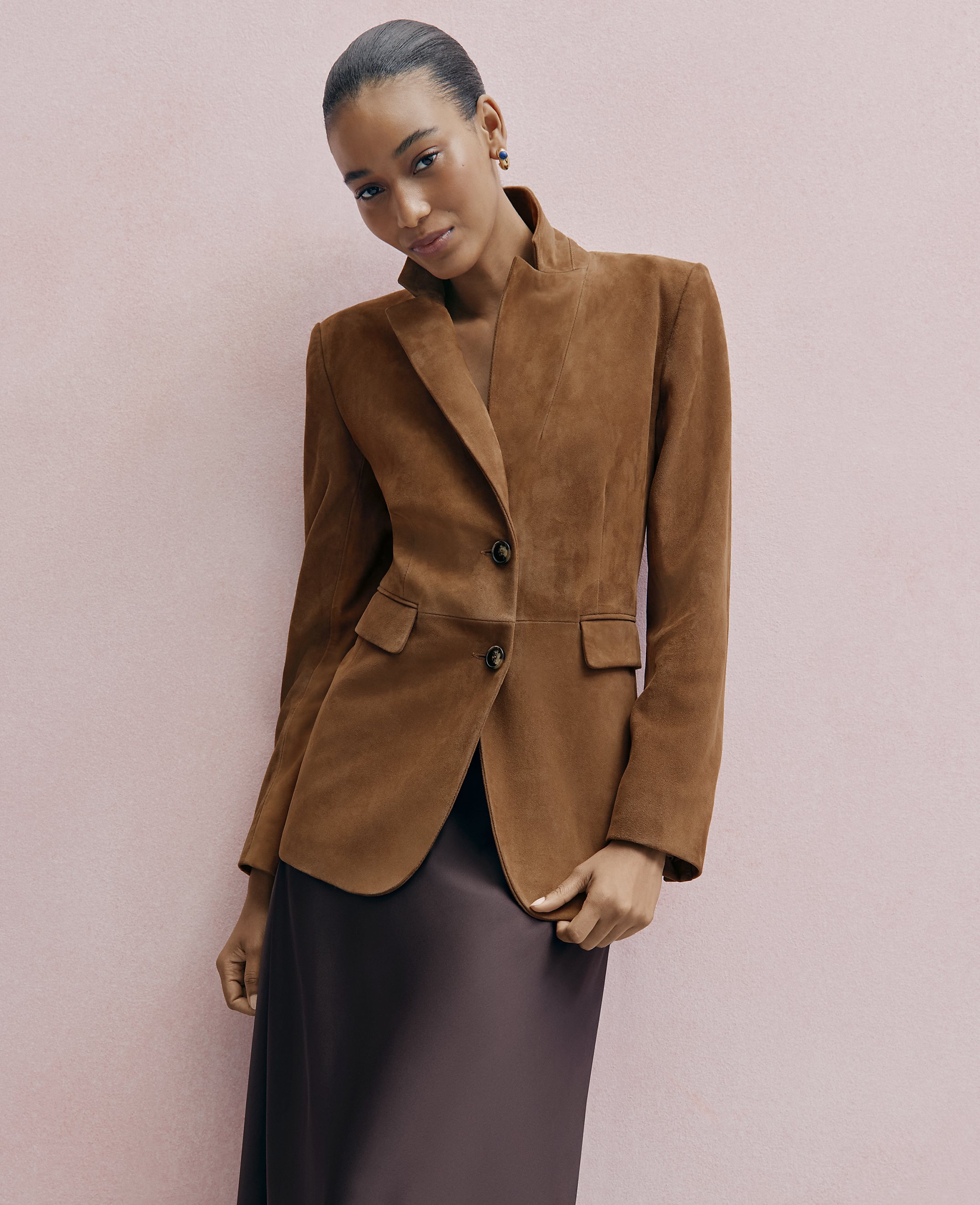 A woman wearing a brown suede blazer and a dark satin skirt poses against a solid light pink background.
