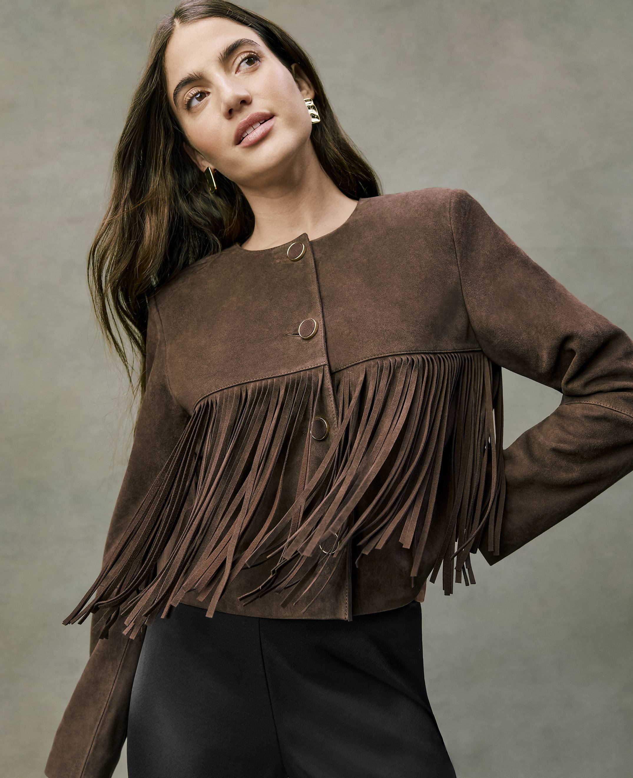A woman wearing a brown suede fringe jacket and black pants poses against a neutral, textured grey background.