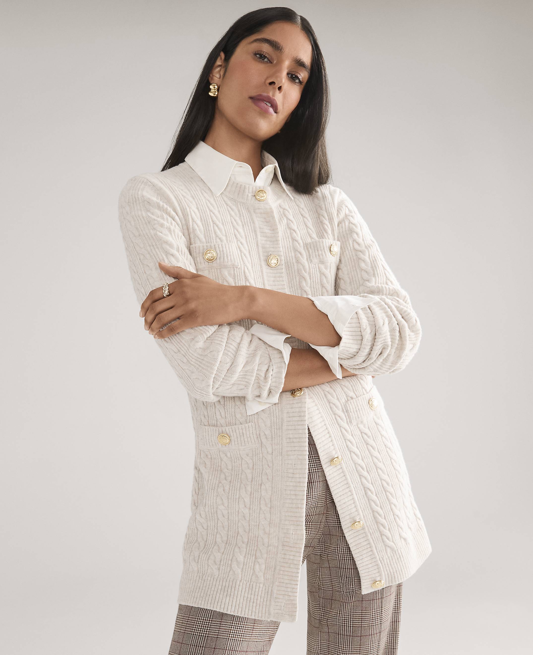 A woman wearing a white cable-knit cardigan over a collared shirt and plaid trousers poses against a plain light gray background.