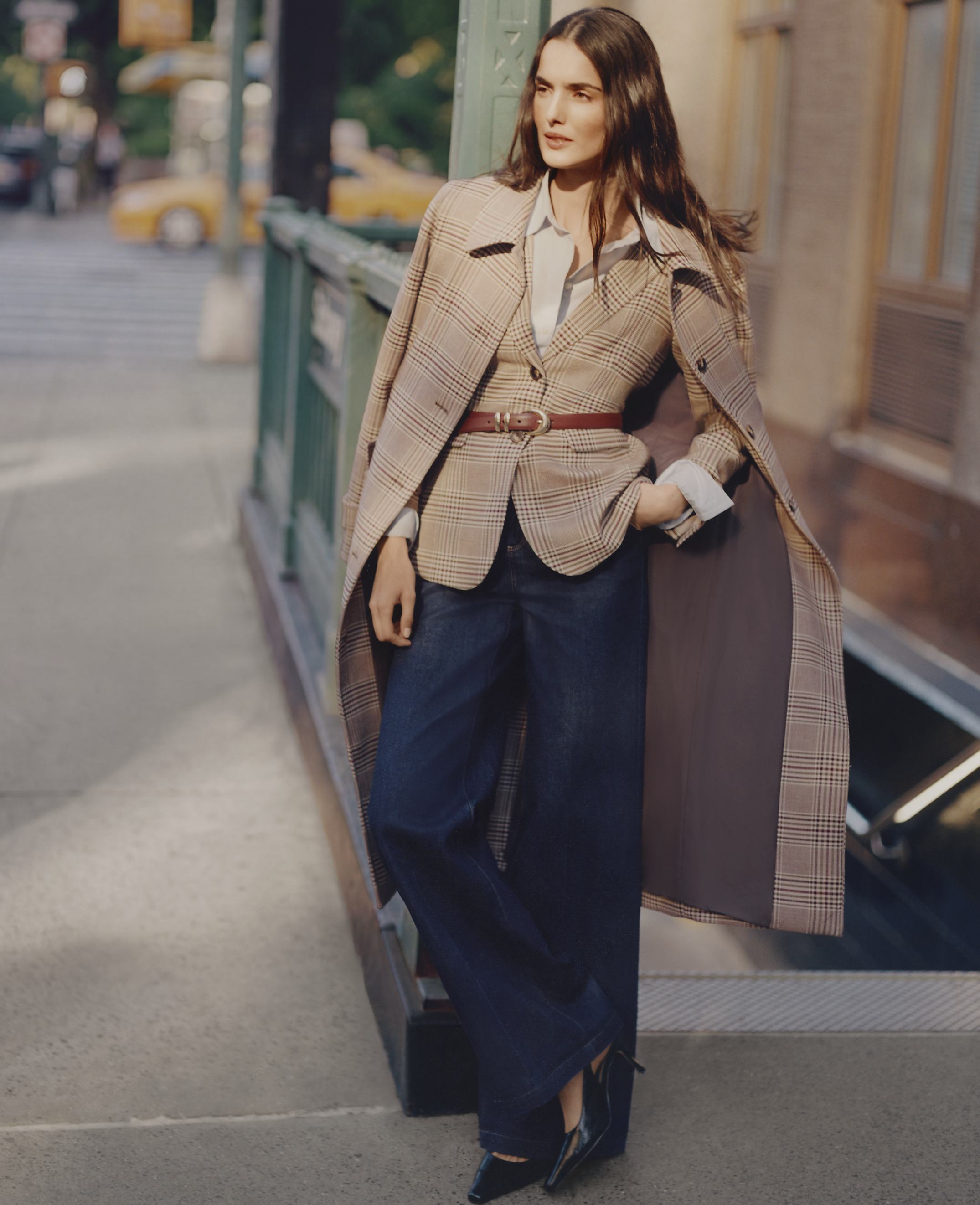 A woman wearing a plaid blazer and wide-leg jeans leans against a green railing on a city sidewalk with blurred yellow taxis in the background.