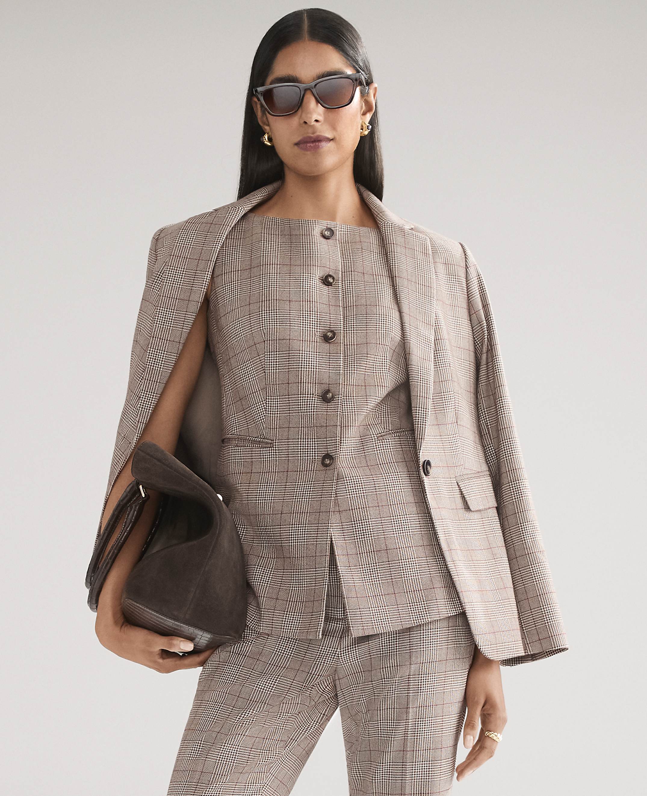 A woman wearing a brown plaid suit and sunglasses holds a dark brown handbag against a plain light gray background.