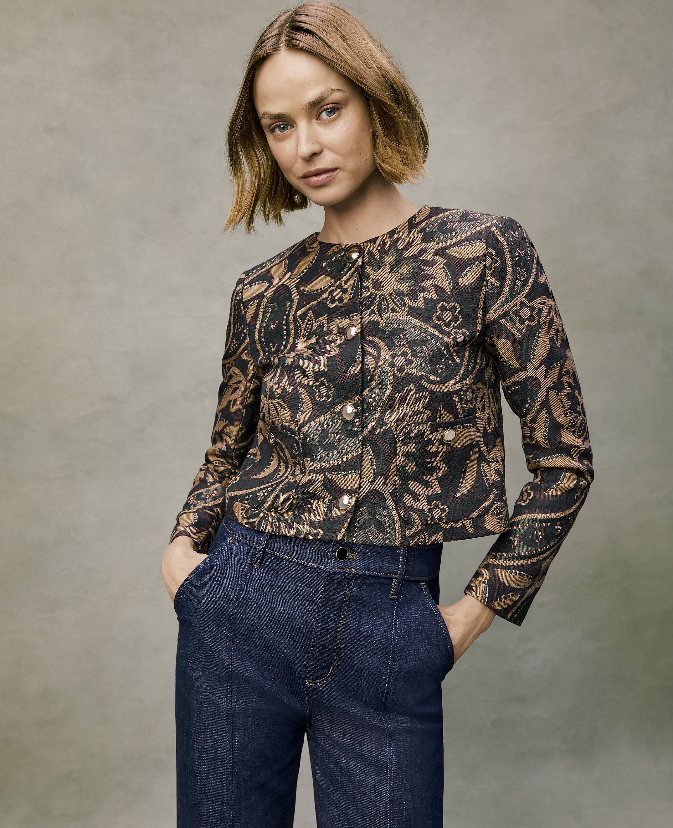 A woman wearing a patterned brown and black jacket and dark blue jeans stands against a neutral, textured grey background.