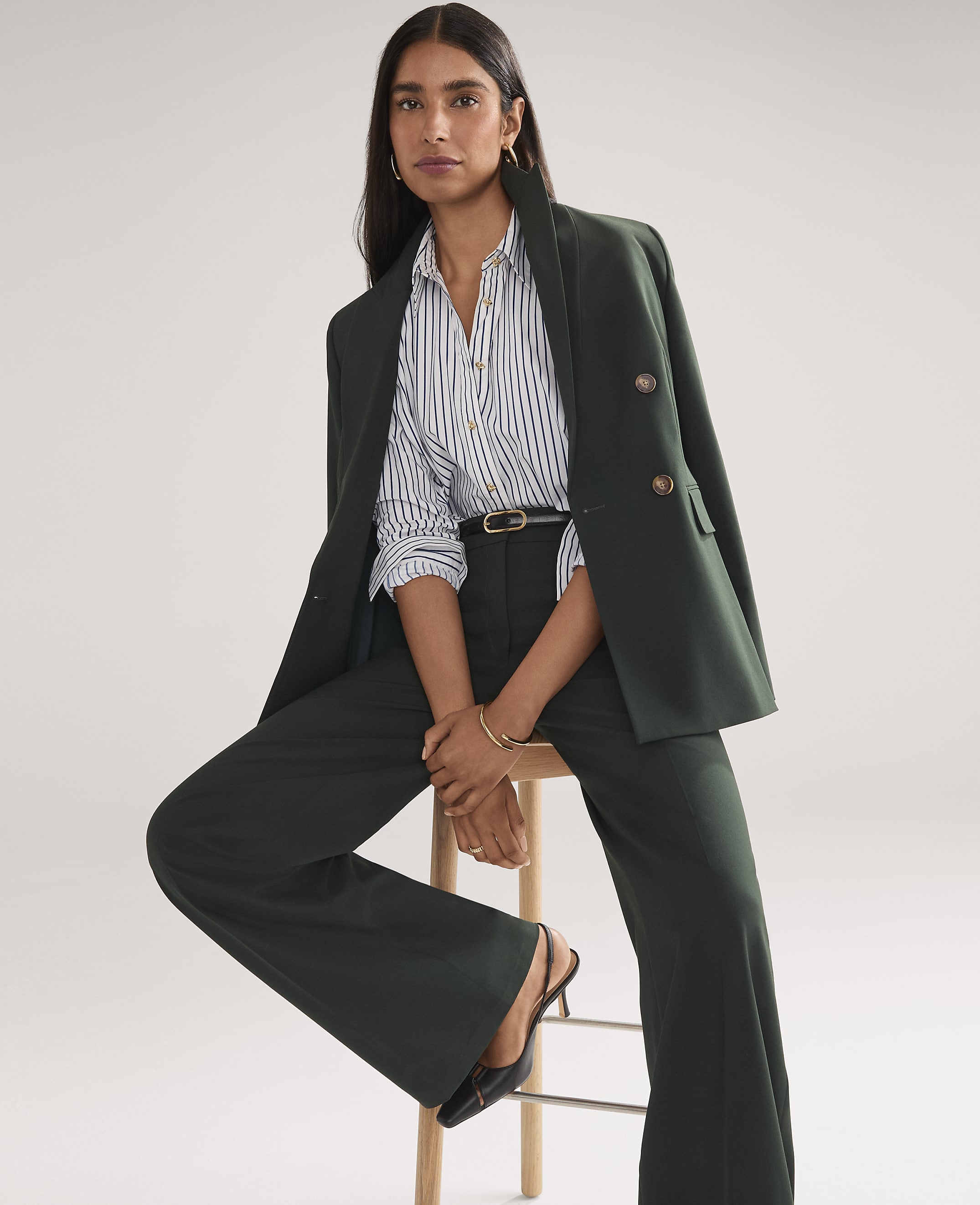 A woman wearing a dark green power suit and a striped shirt stands against a plain white background.