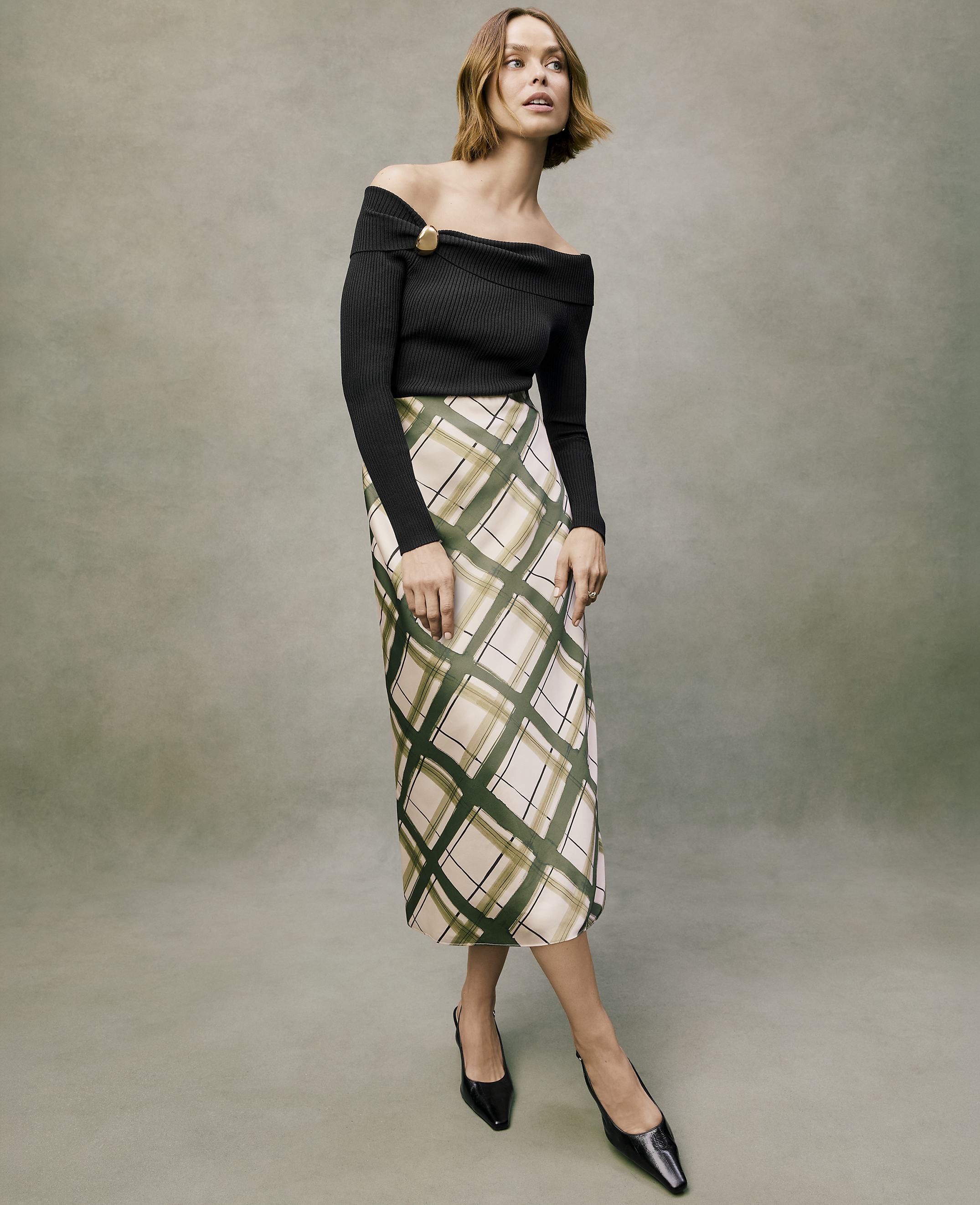 A woman wearing a black off-the-shoulder top and a plaid midi skirt stands against a muted green textured background.