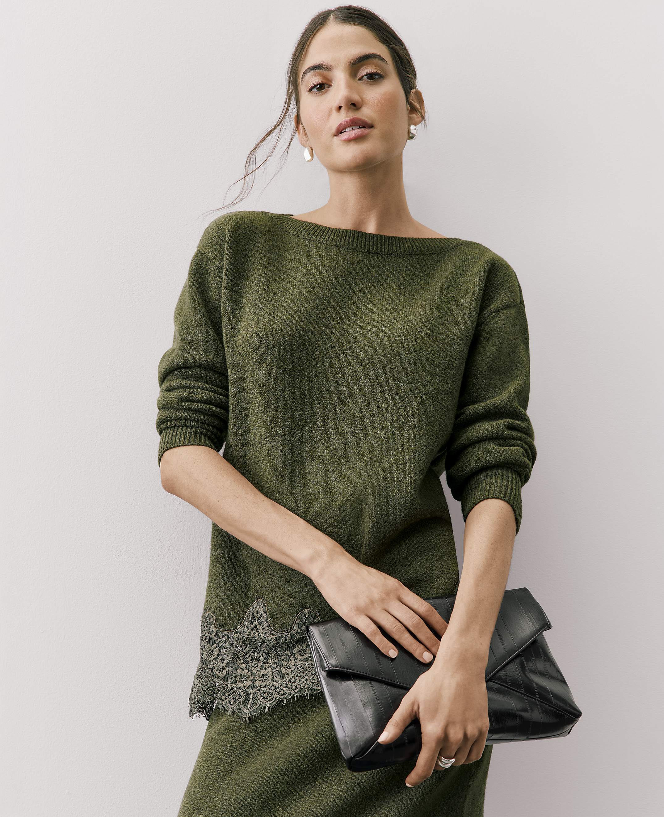 A woman wearing an olive green sweater with lace detailing and holding a black clutch stands against a plain white background.