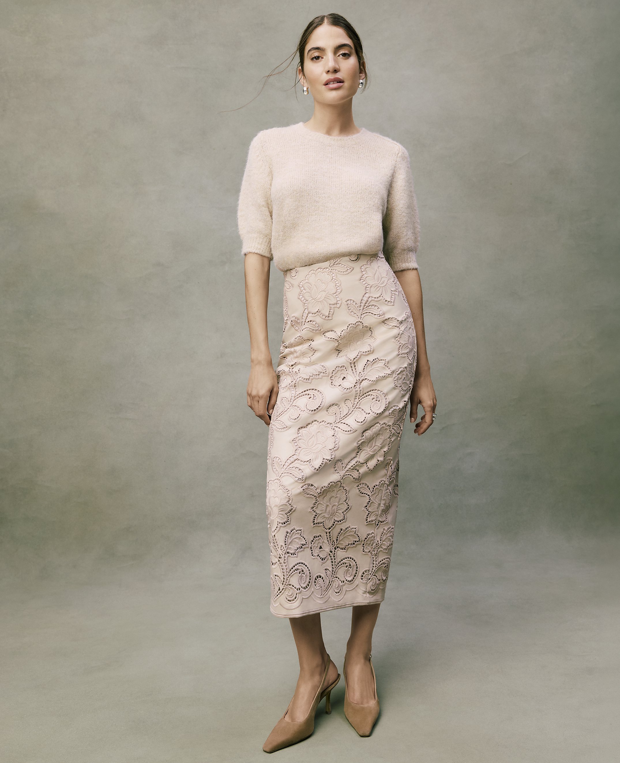 A woman wearing a fuzzy cream sweater, an embroidered pale pink skirt, and nude heels stands against a textured grey background.