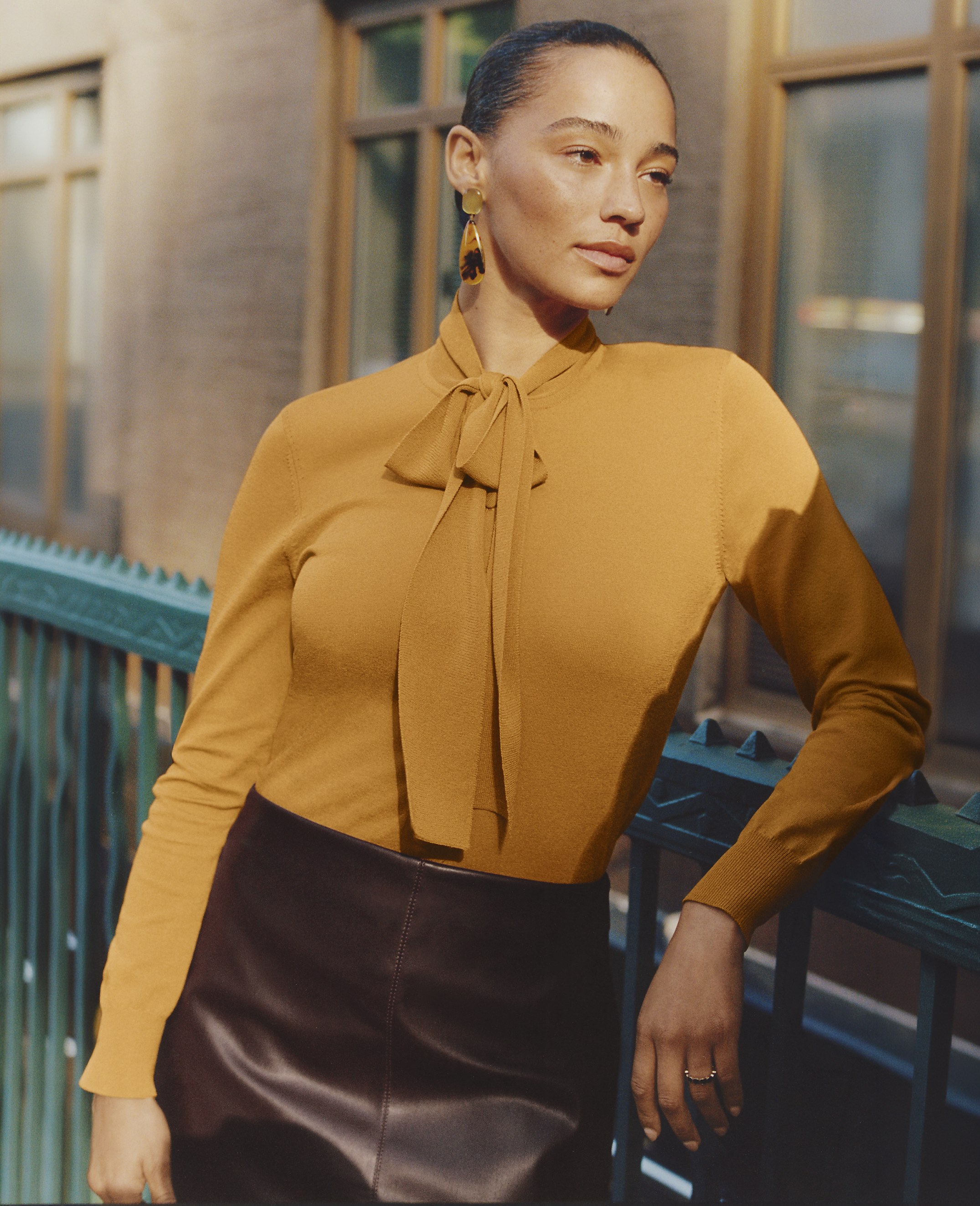 A woman wearing a mustard-yellow pussy-bow blouse and a dark leather skirt leans against a teal railing in front of a blurred city building.