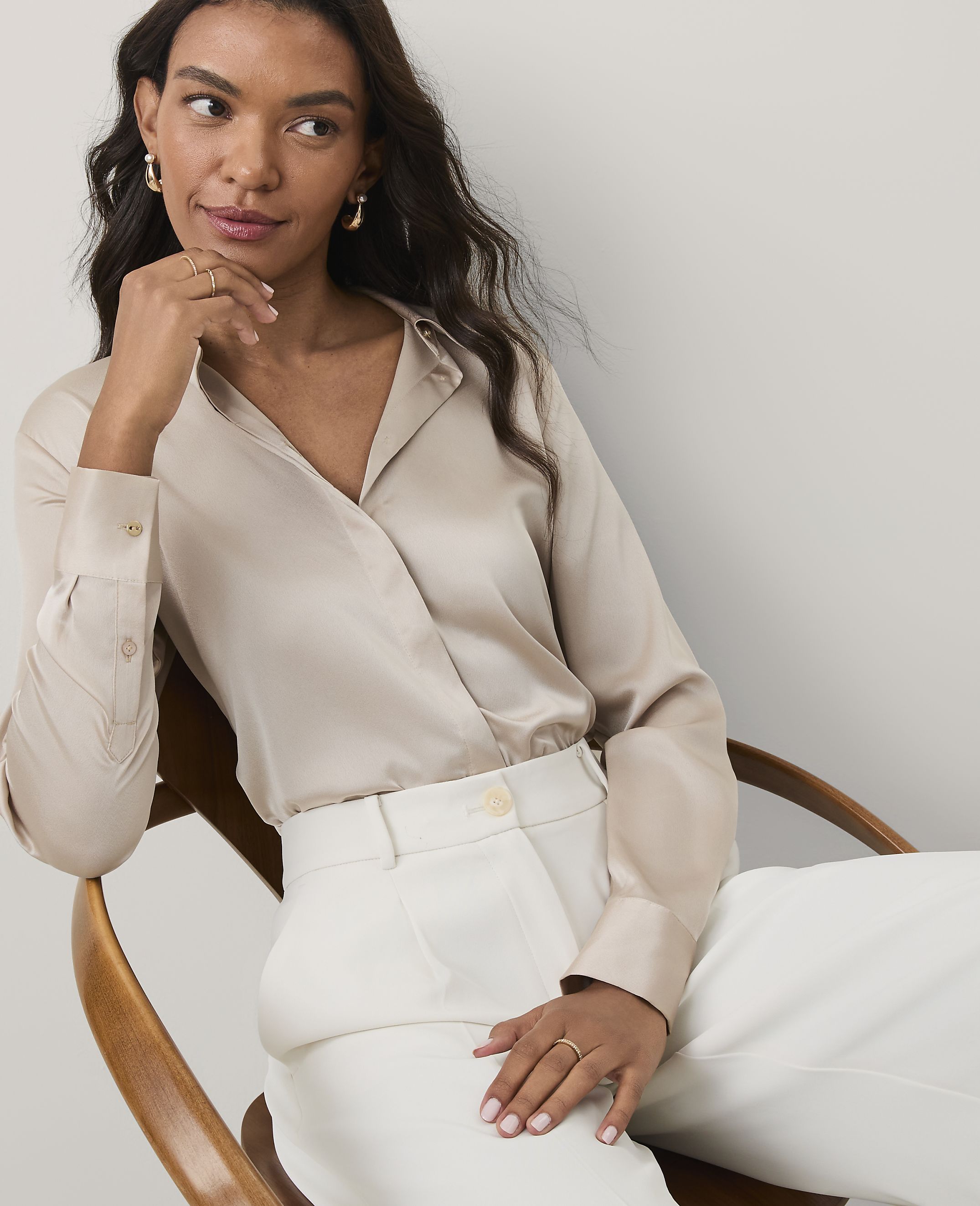 A woman wearing a champagne-colored satin blouse and white trousers sits in a wooden chair against a plain light gray background.