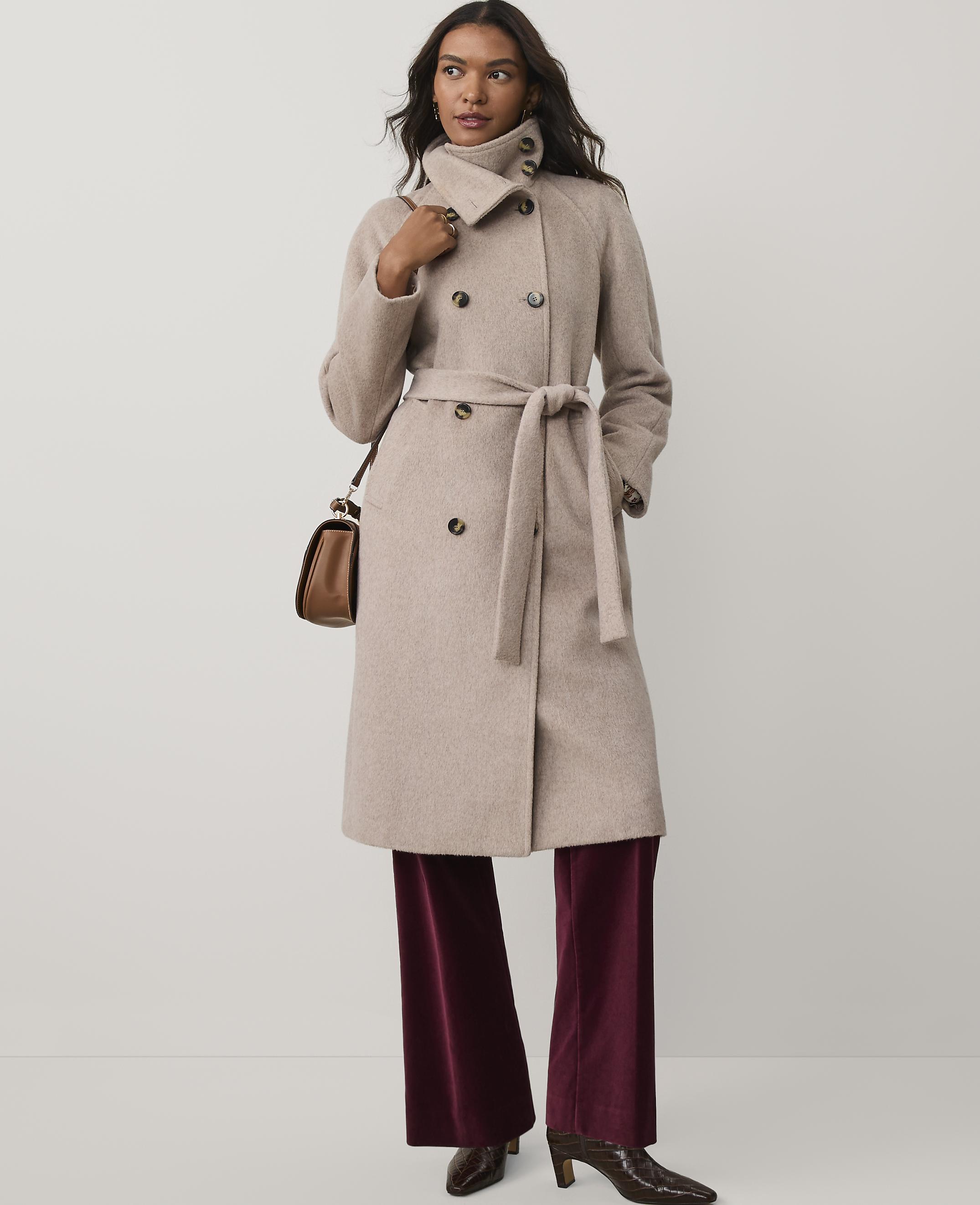 A woman wearing a beige belted coat and burgundy velvet trousers stands against a plain white background.