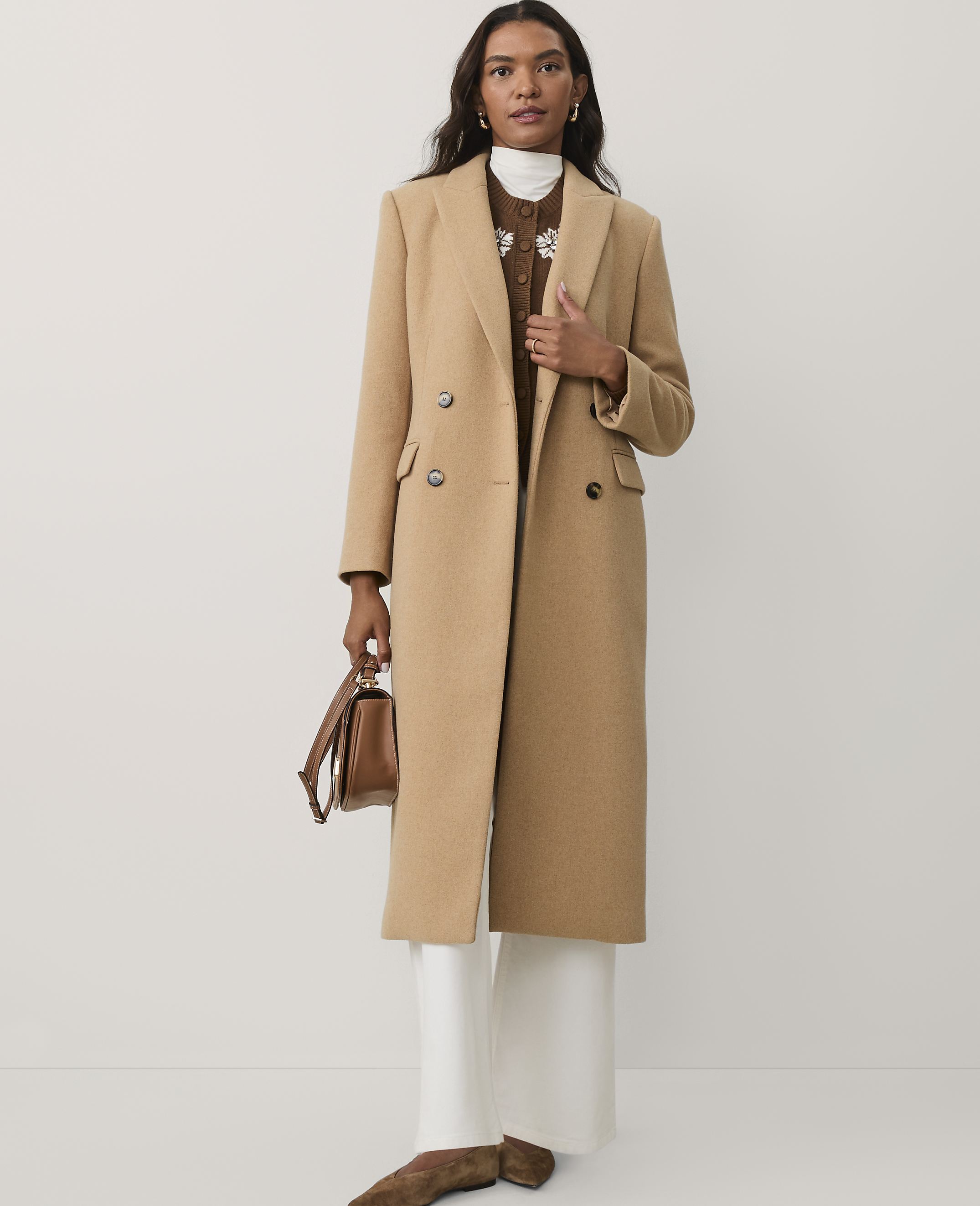 A woman wearing a long camel-colored coat, white trousers, and brown shoes stands against a plain white background.