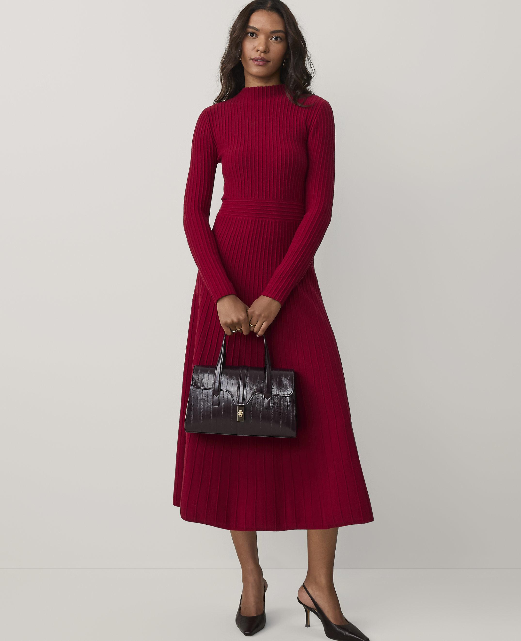 A woman wearing a long-sleeved red ribbed dress and black heels holds a dark burgundy handbag against a plain white background.