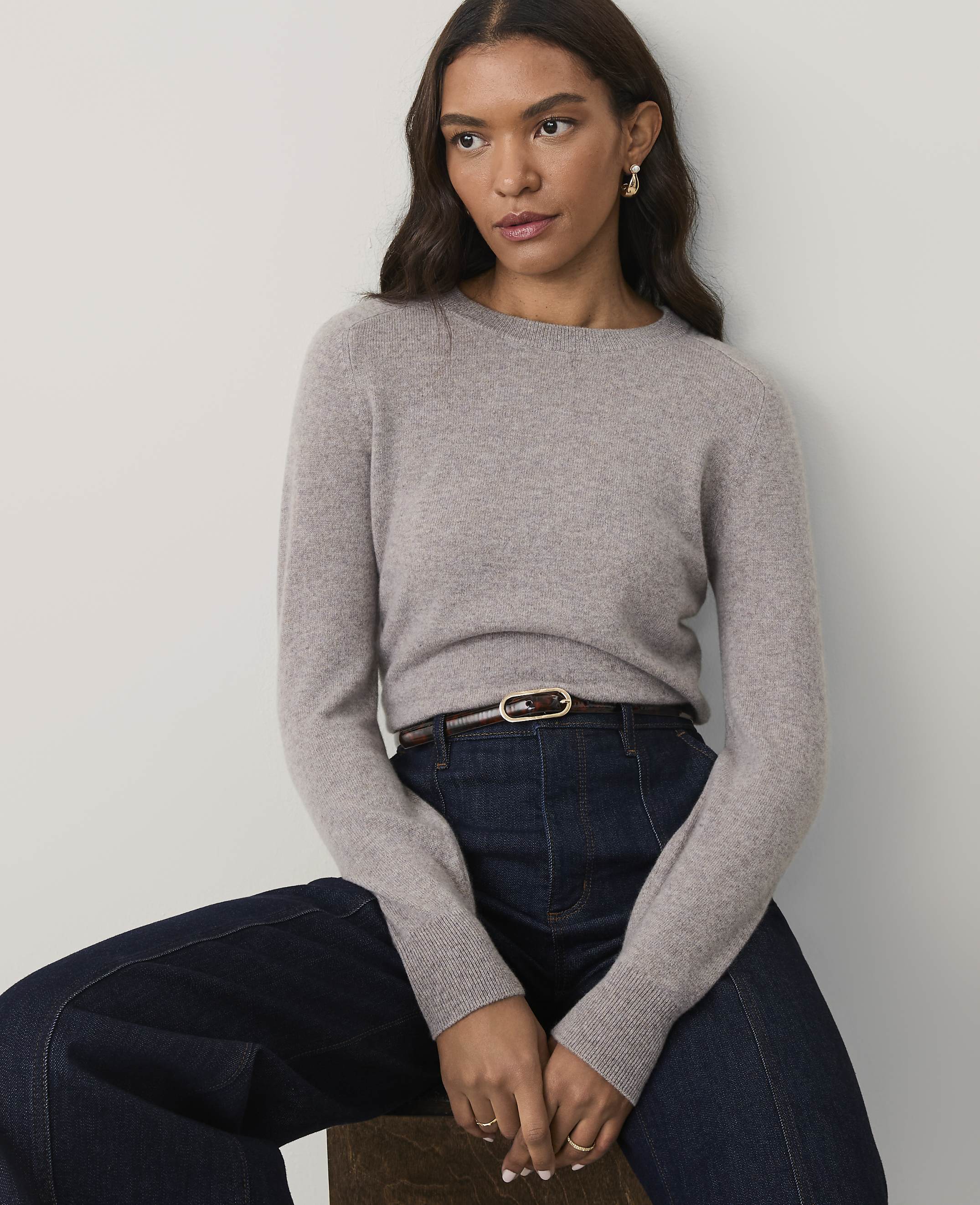 A woman wearing a grey sweater and dark blue jeans sits against a plain white background.