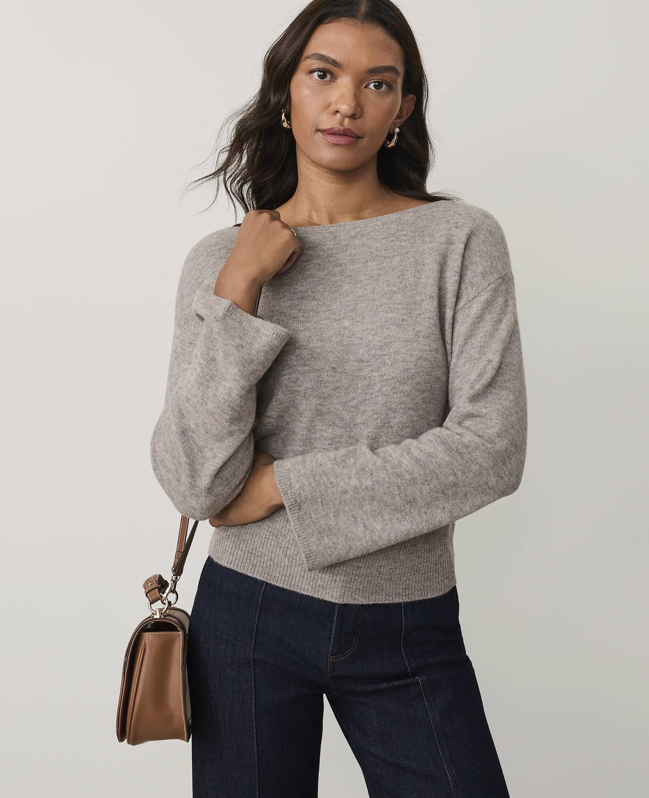 A woman wearing a beige sweater and dark blue jeans stands against a plain light gray background.