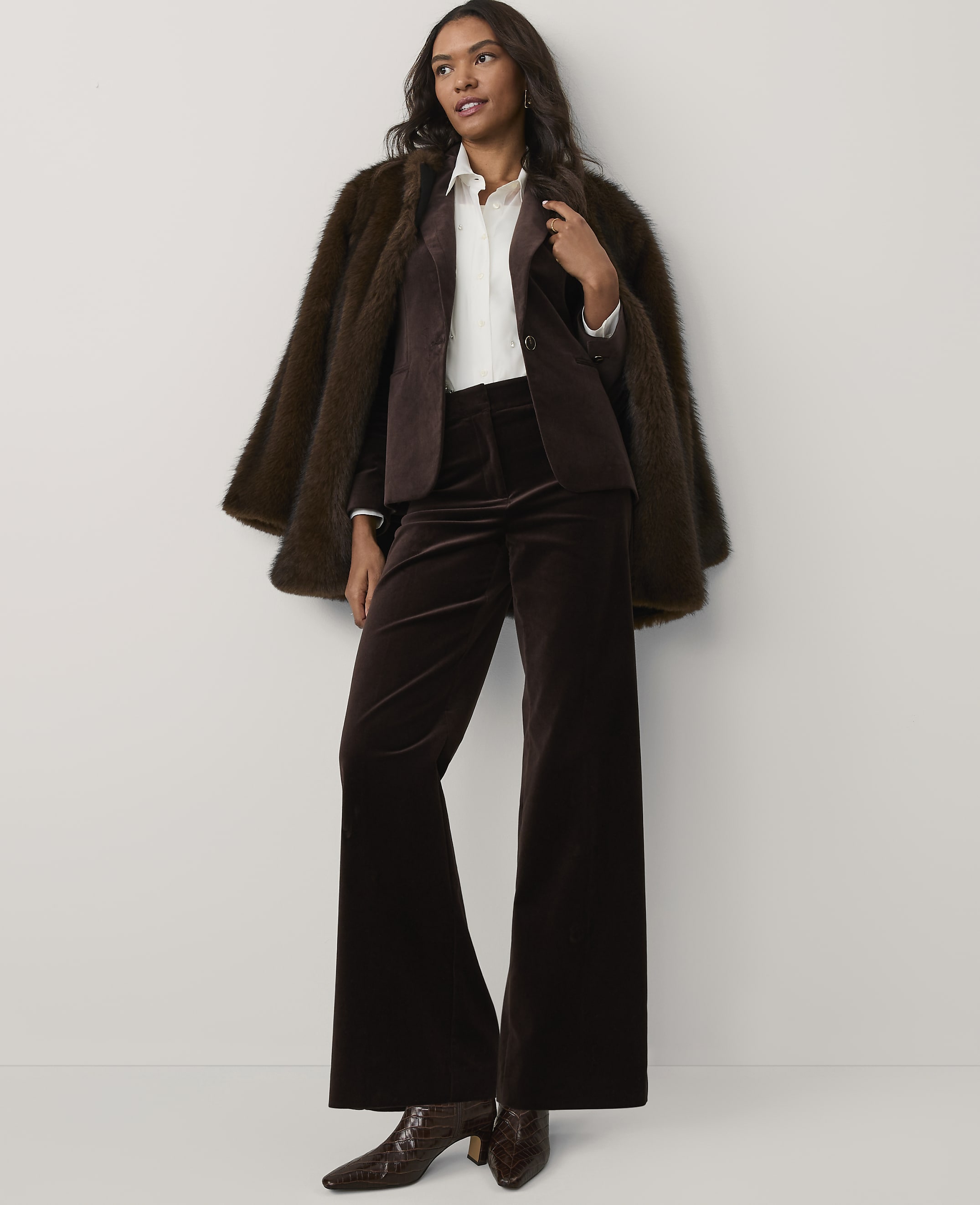A woman wearing a brown faux-fur coat, blazer, and wide-leg velvet trousers stands against a plain white background.