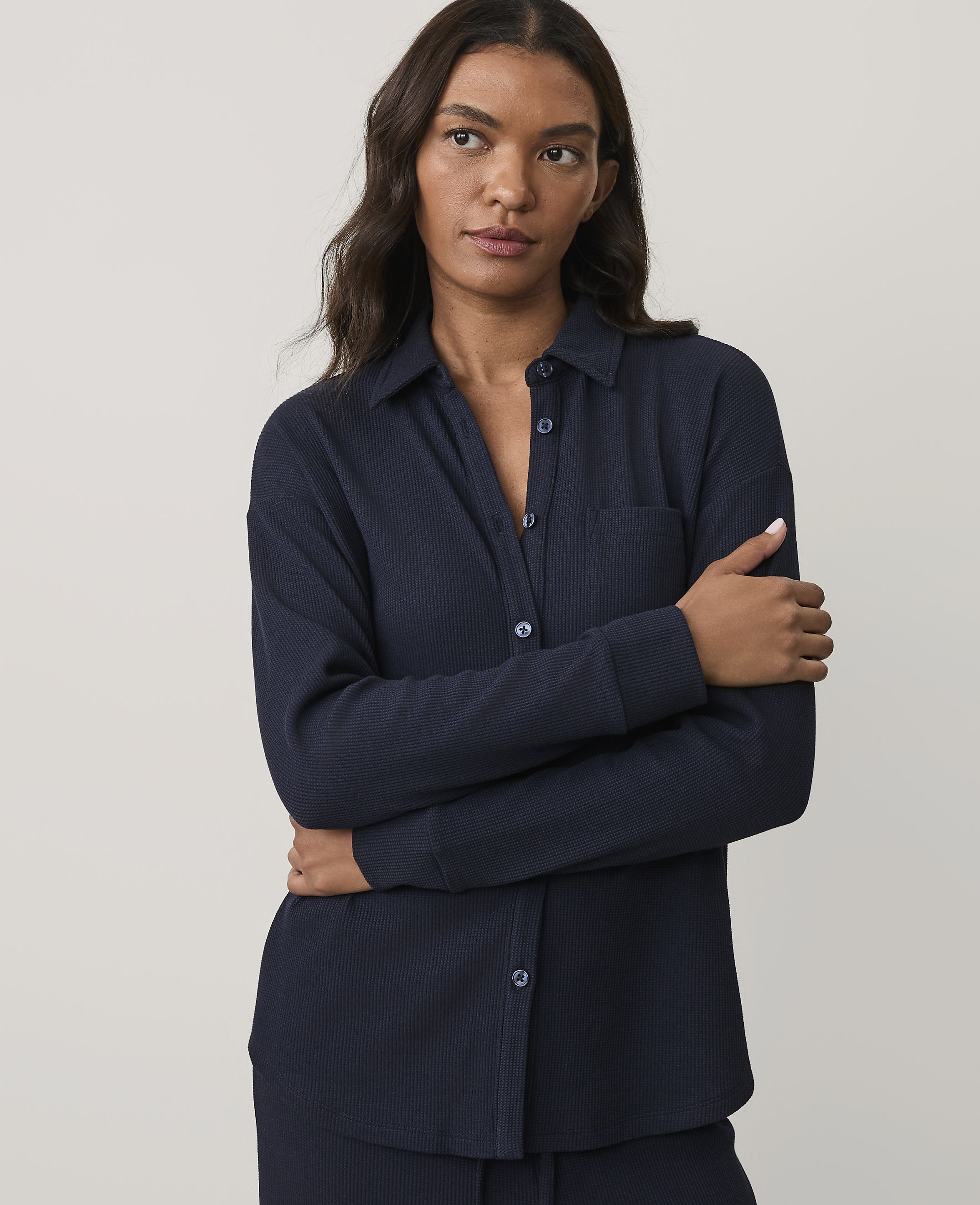 A woman wearing a navy blue ribbed button-down shirt stands with her arms crossed against a plain, light grey background.