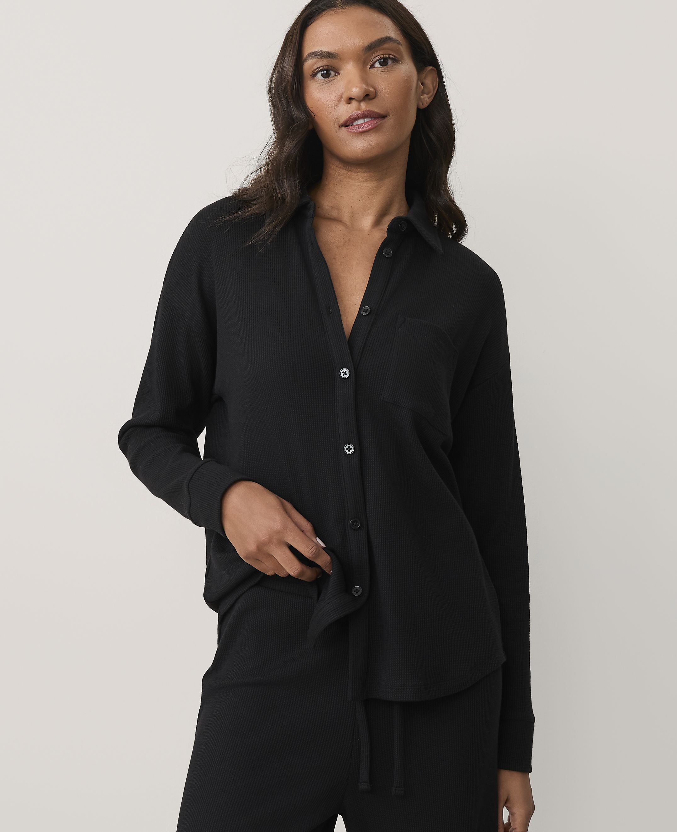 A woman wearing a black ribbed pajama set stands against a plain, light-colored background.