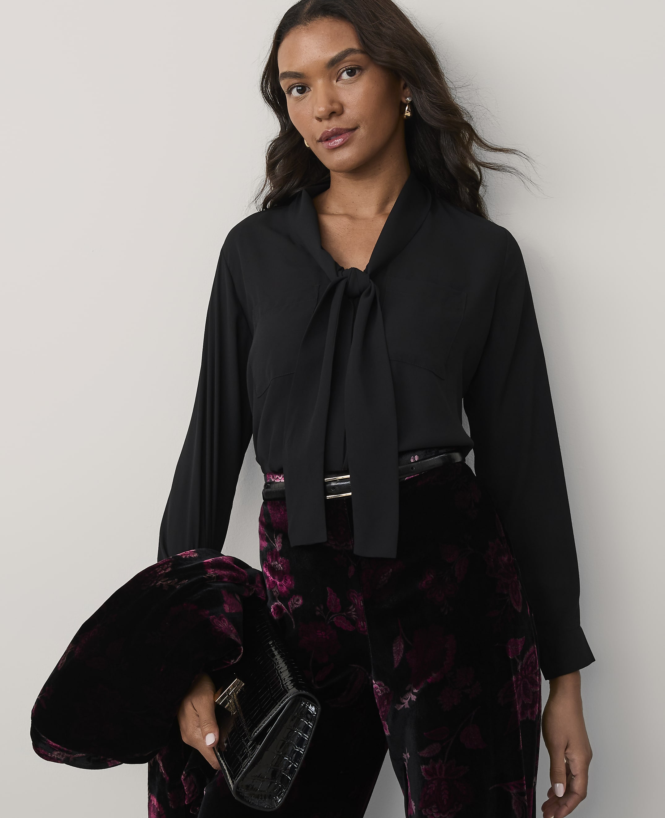 A woman wearing a black tie-neck blouse and floral velvet trousers stands against a plain, light-colored background.