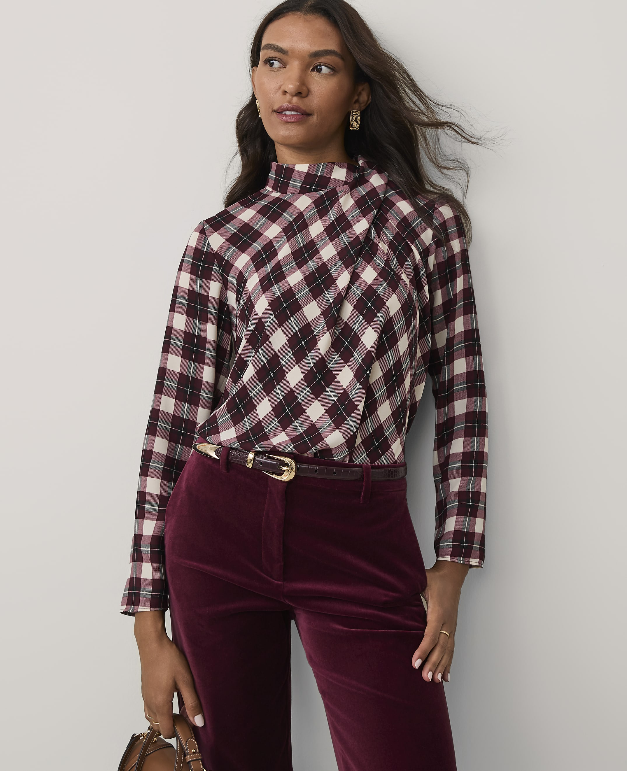 A woman wearing a plaid top and burgundy velvet trousers stands against a plain light gray background.