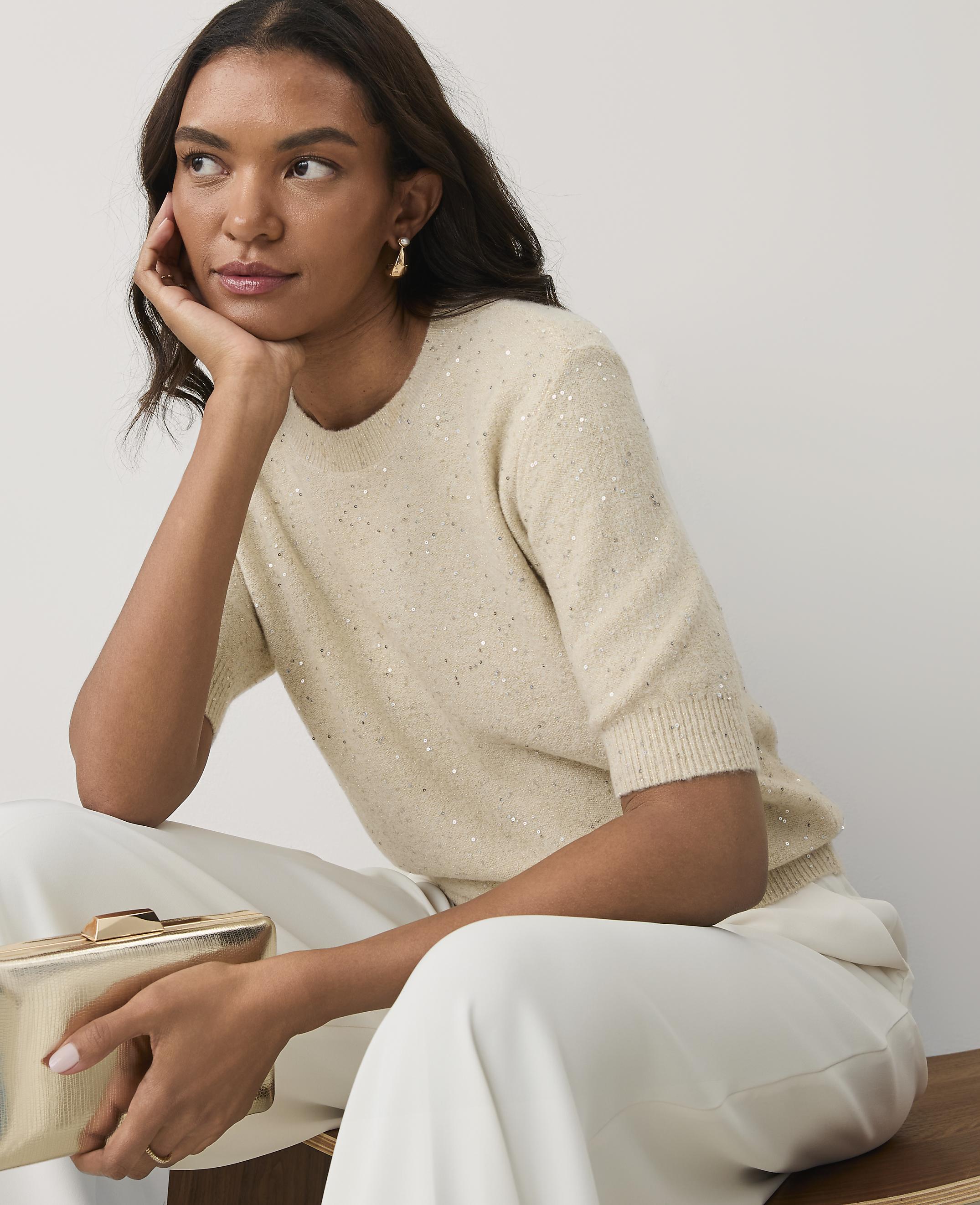 A woman in a shimmering cream sweater and white trousers holds a gold clutch against a plain light gray background.