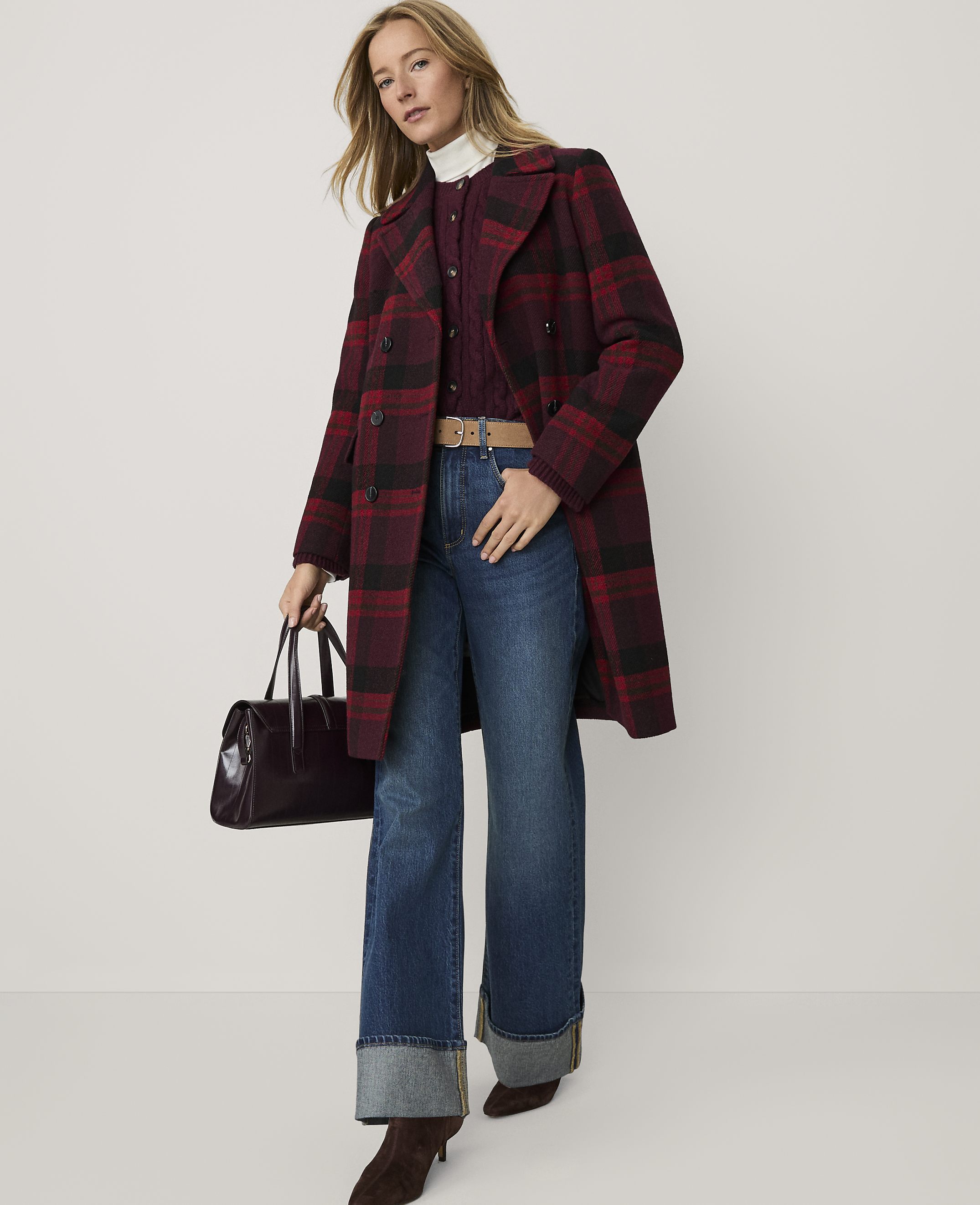 A woman wearing a red plaid coat, blue jeans, and brown boots stands against a plain white background while holding a dark handbag.