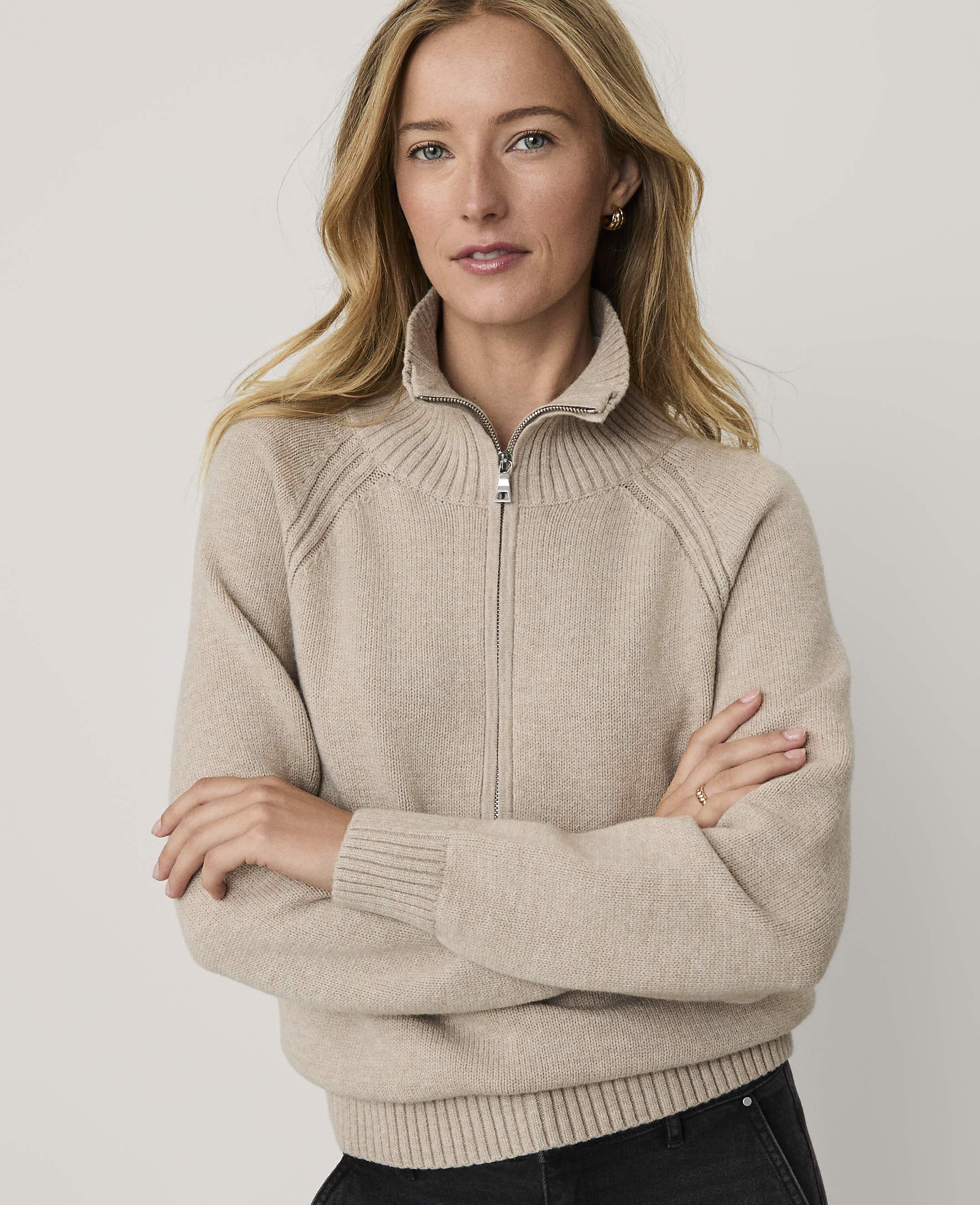 A woman with blonde hair wearing a beige zip-up sweater stands with her arms crossed against a plain, light-colored background.
