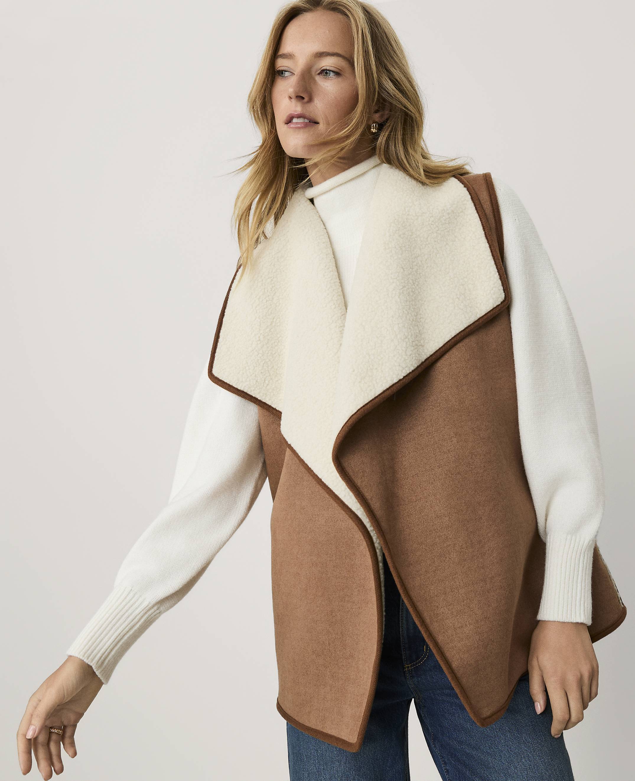 A woman wearing a brown and cream vest over a white sweater and blue jeans stands against a plain, light-colored background.
