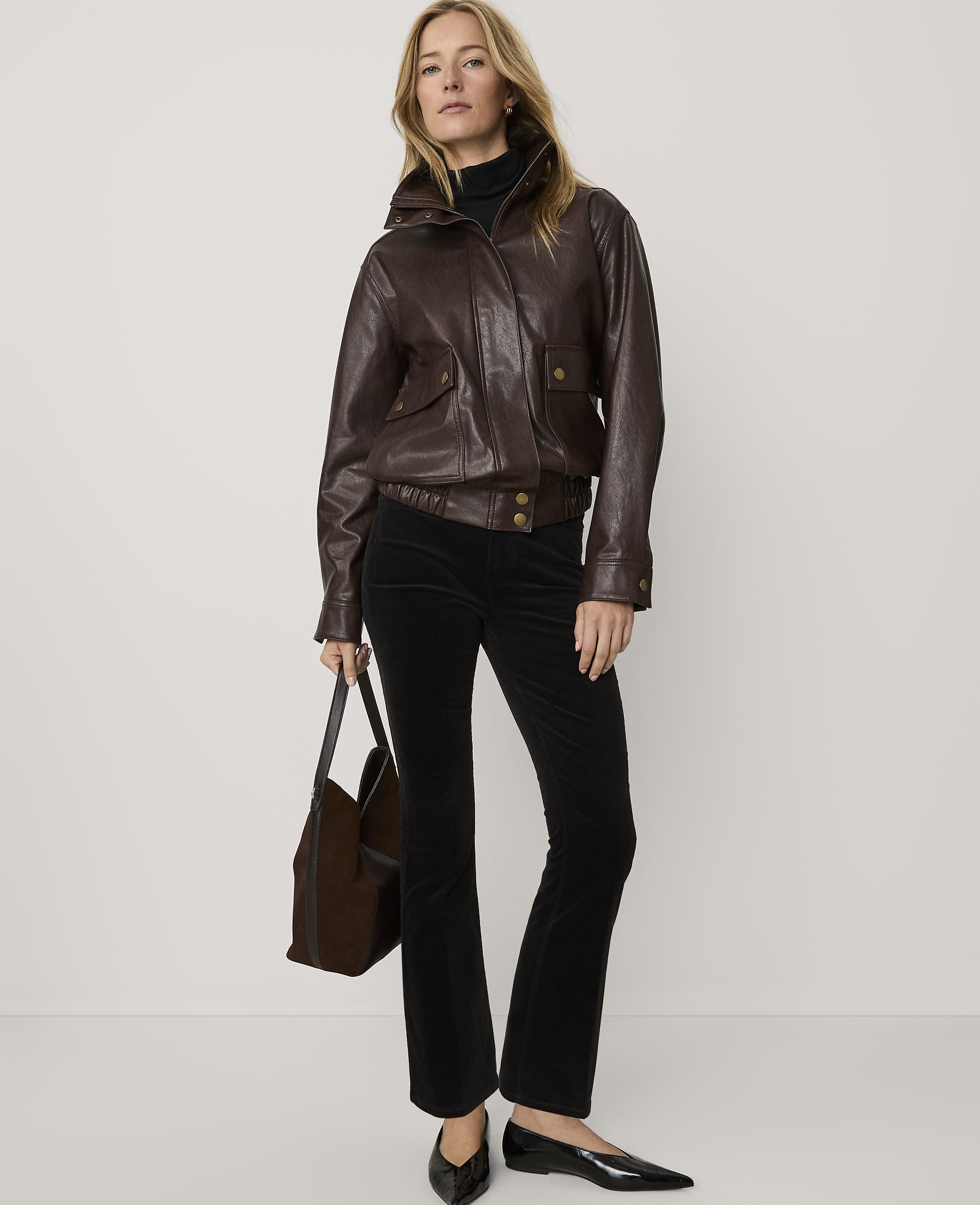 A woman wearing a brown leather jacket, black trousers, and black heels stands against a plain white background while holding a brown handbag.