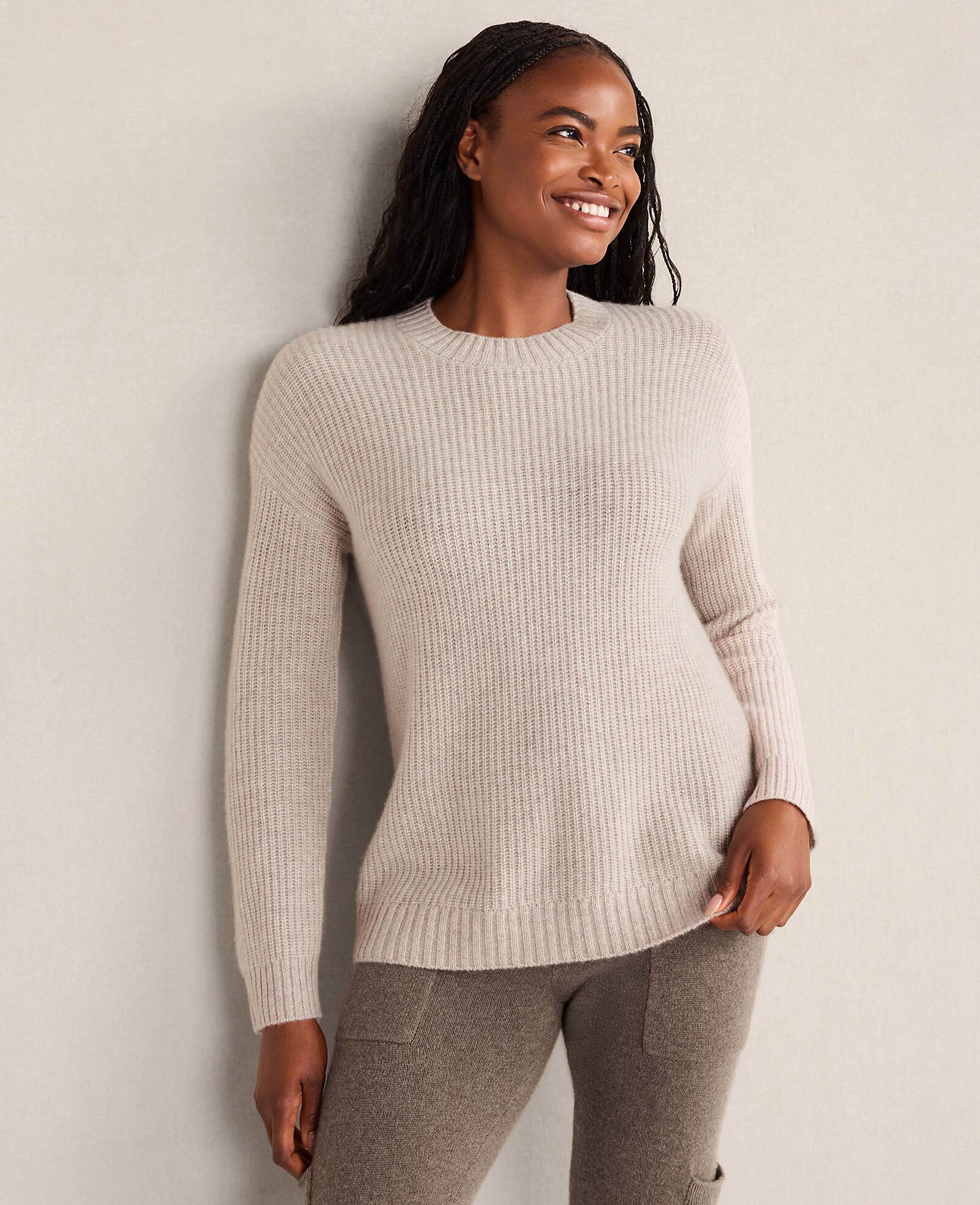 A smiling woman wearing a light beige ribbed sweater and brown pants leans against a plain, neutral-colored wall.