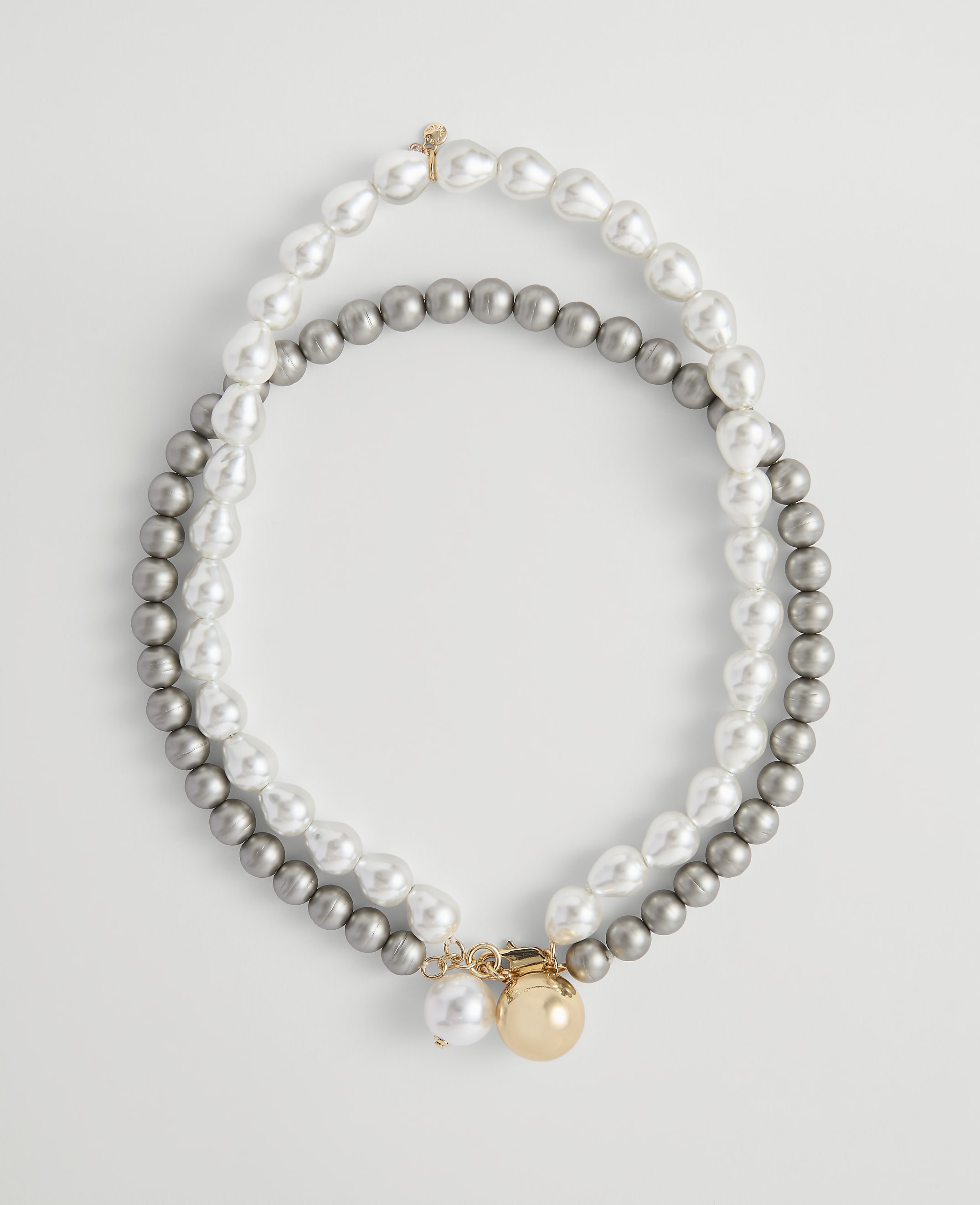 Two strands of pearls, one white and baroque and the other grey and round, are laid out in an oval shape against a plain white background.