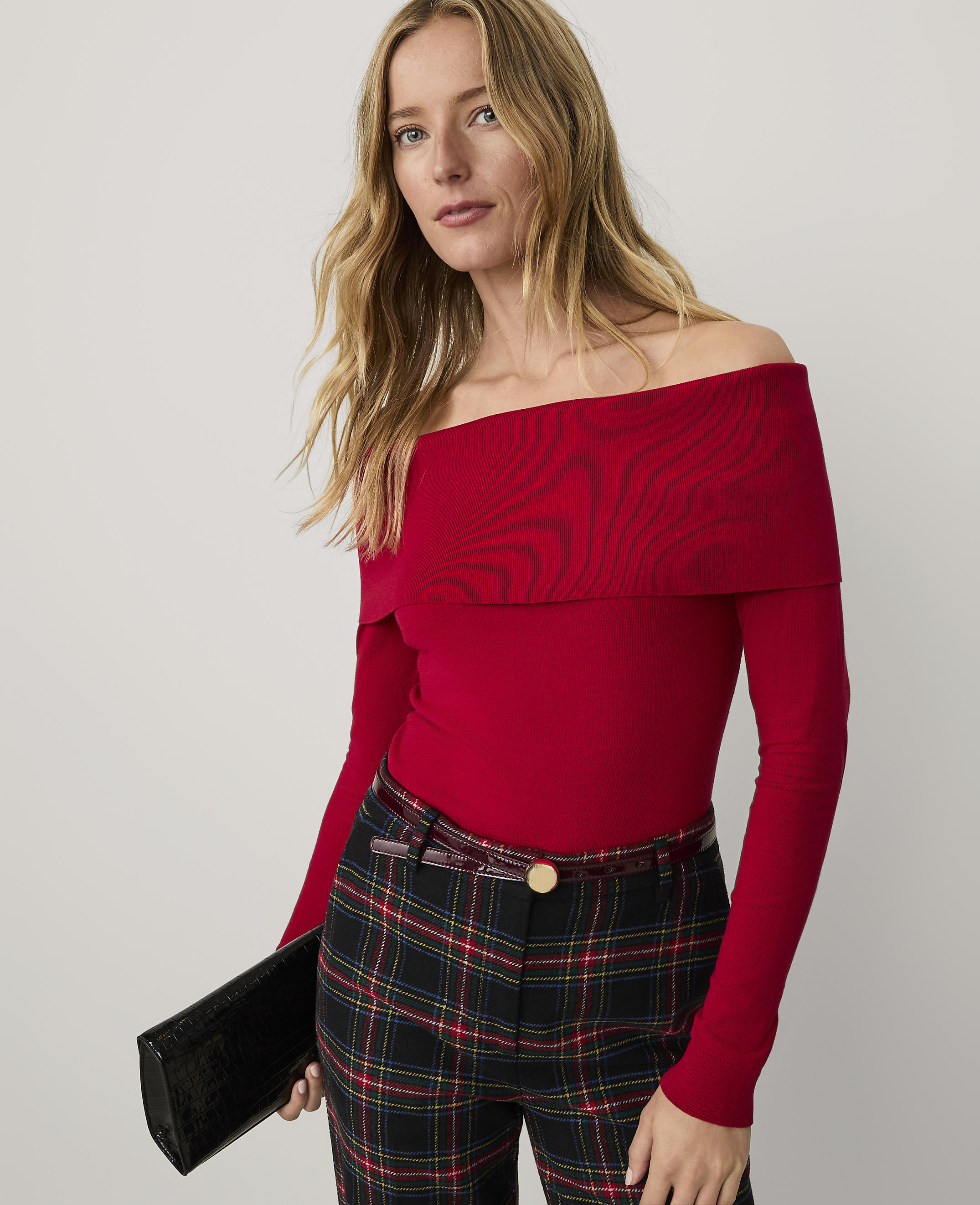 A woman wearing a red off-the-shoulder top and plaid trousers stands against a plain white background.