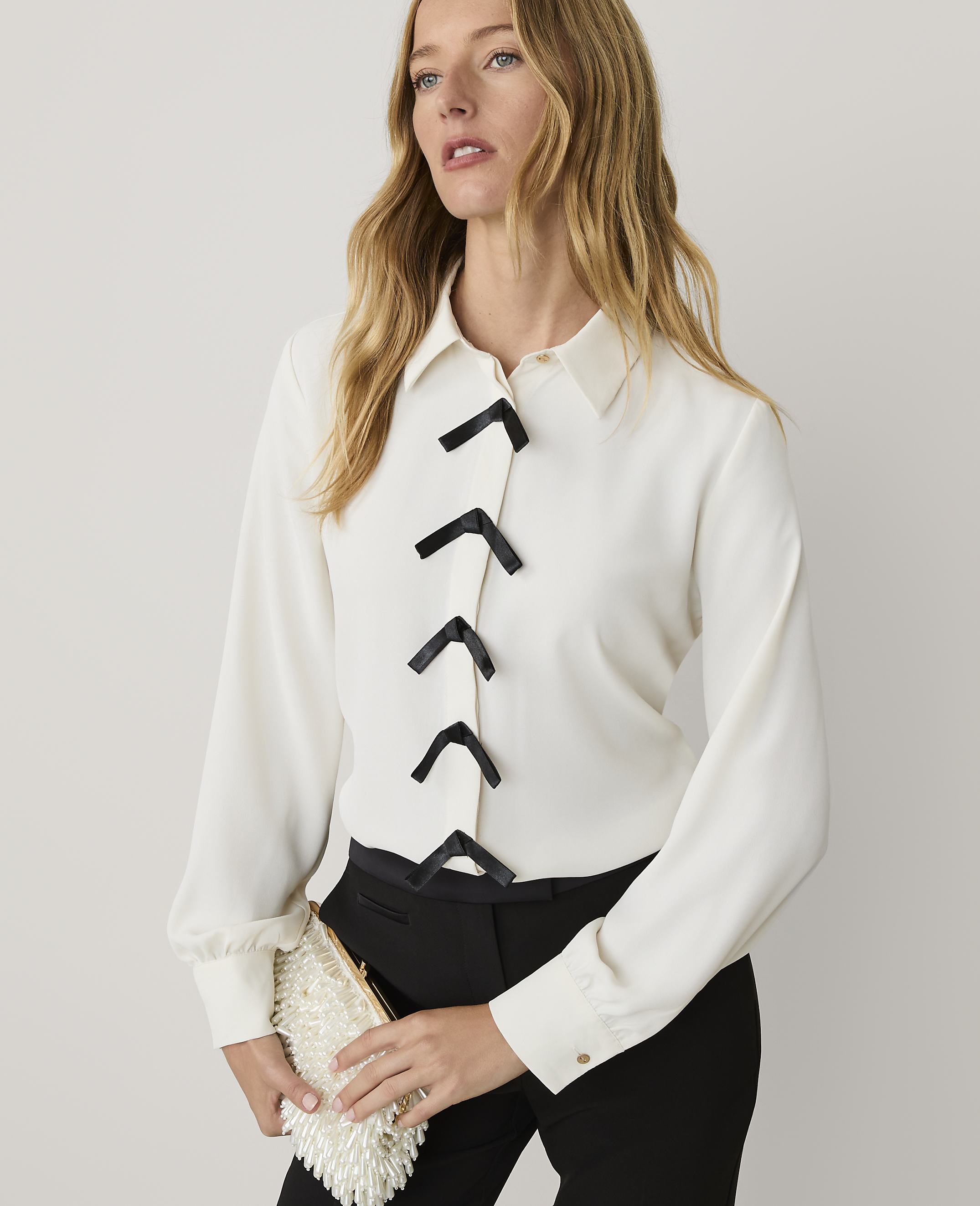 A blonde woman wearing a white blouse with black accents and black trousers stands against a plain white background.