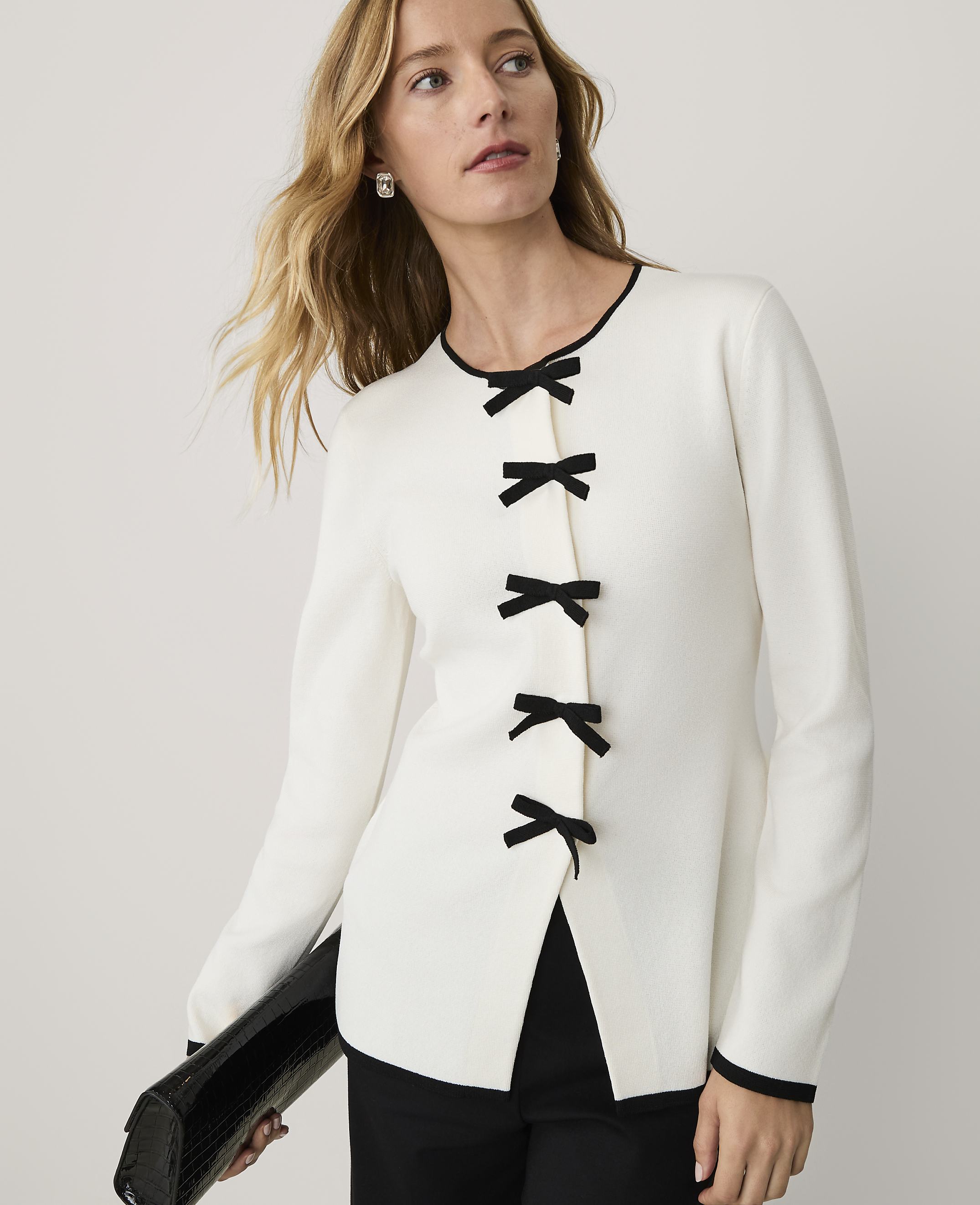 A woman wearing a white long-sleeved top with black bow closures and black trousers stands against a plain white background.