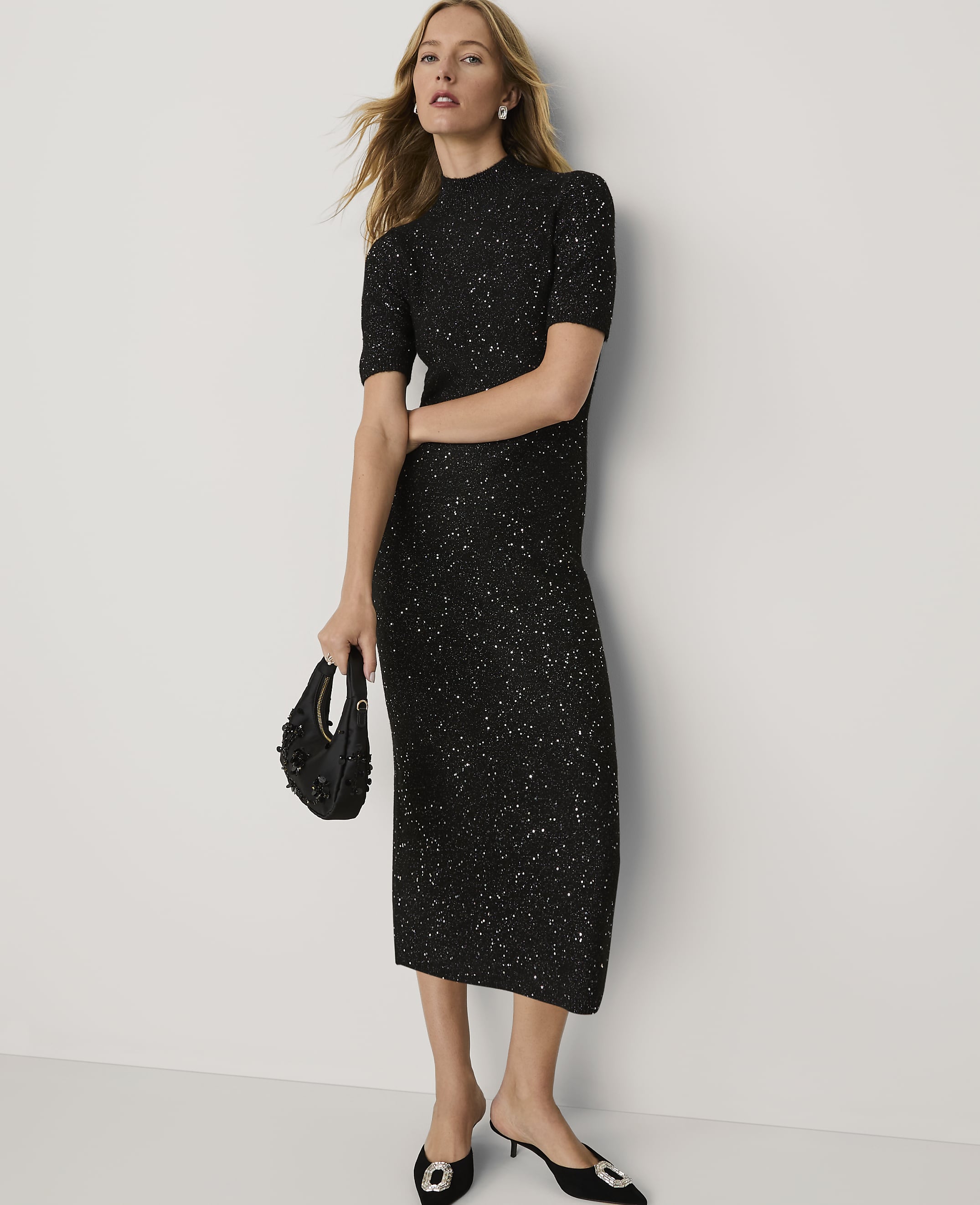 A woman wearing a sparkling black midi dress and black heels stands against a plain white background.